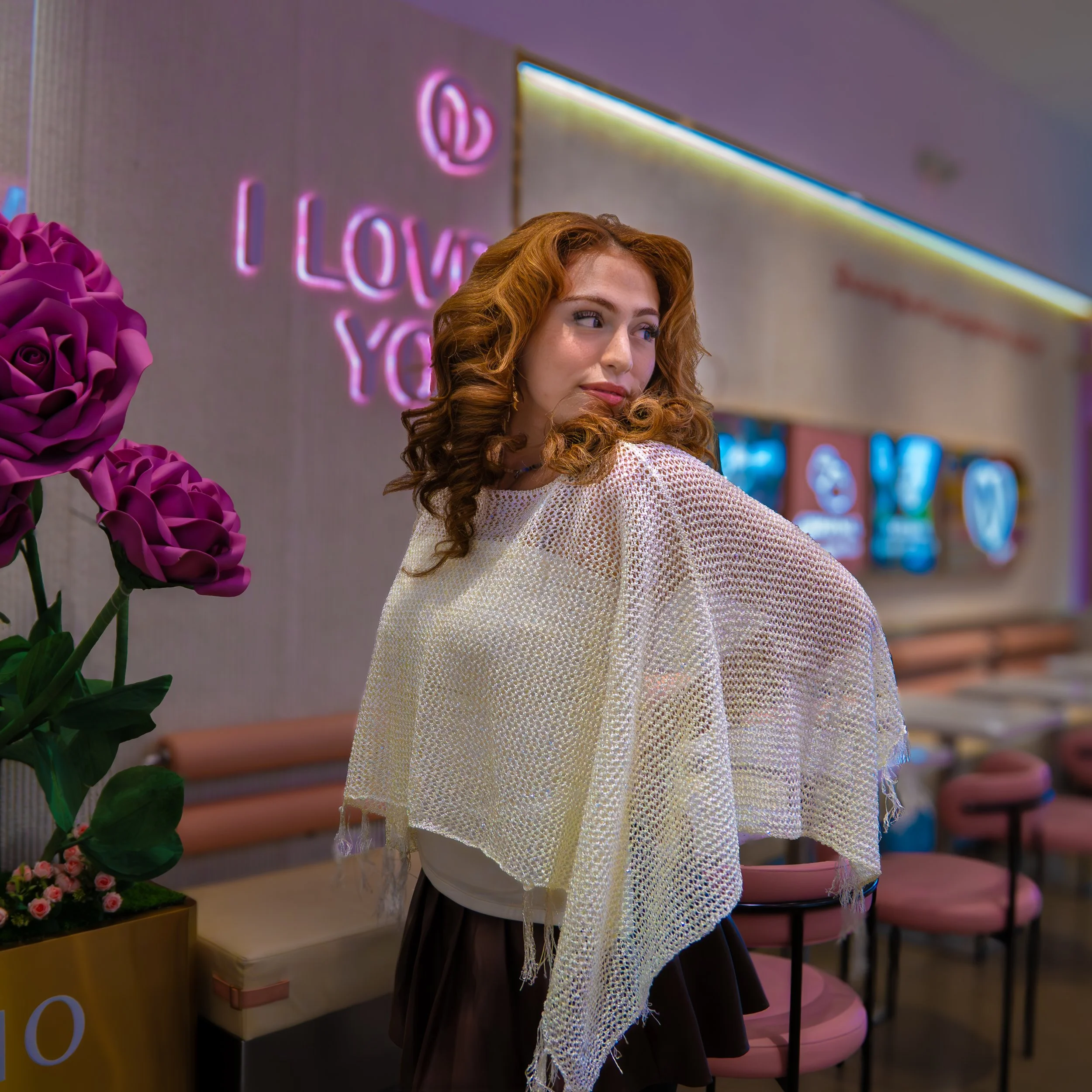 A woman with long, curly red hair looking to the side in front of a floral display and neon sign that says 'Love You' in pink lights.