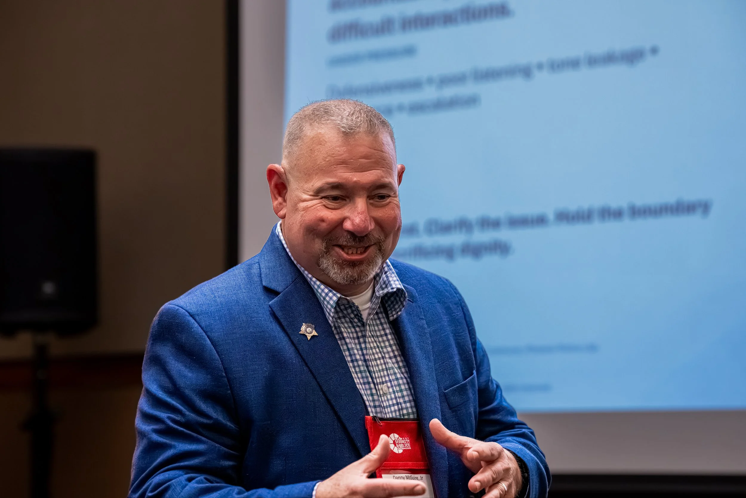 Smiling man with short gray hair and a beard, wearing a blue blazer and checkered shirt, standing in front of a screen with text, holding a red folder or badge, possibly giving a presentation.