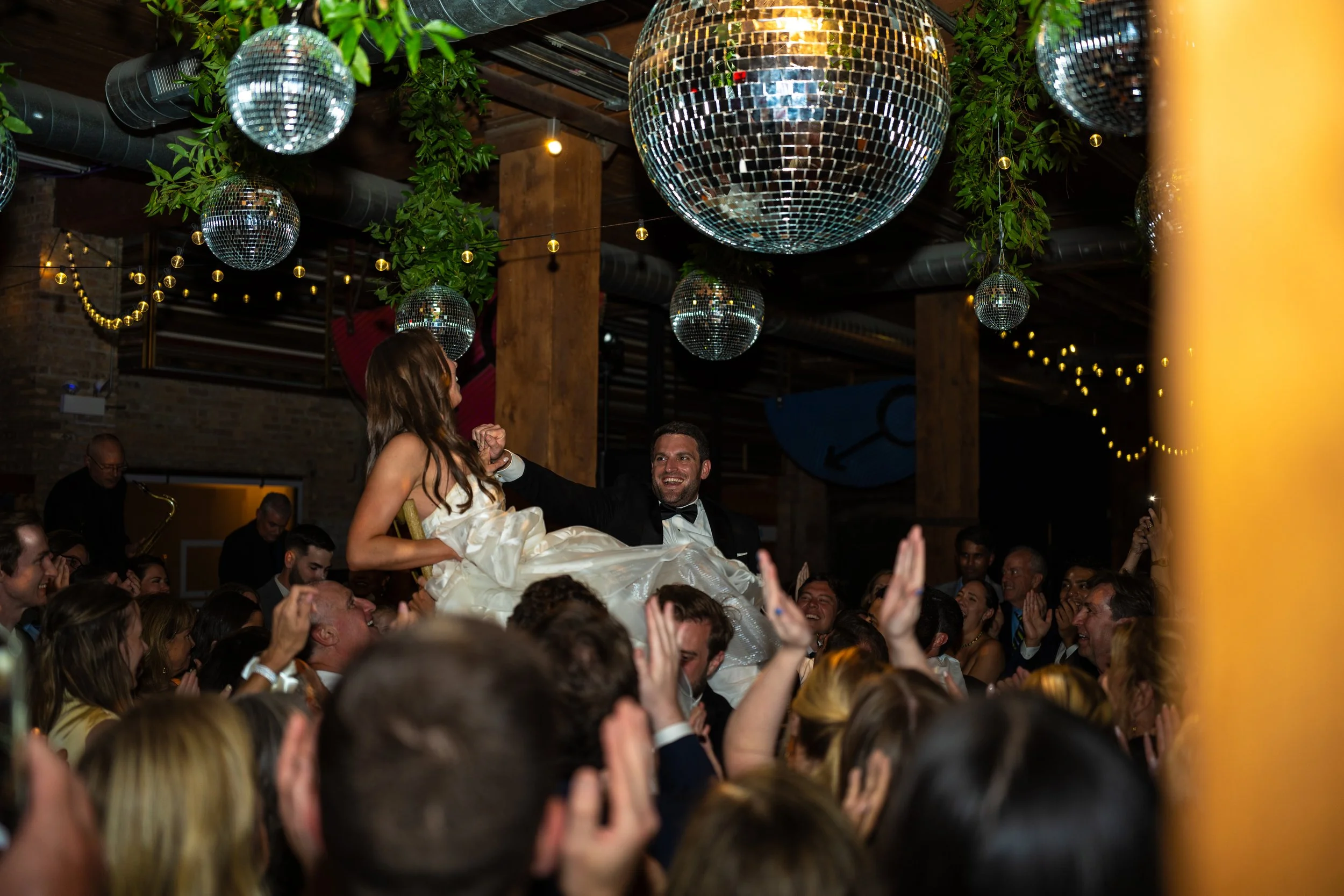 A wedding celebration with a bride and groom dancing on a crowded dance floor, surrounded by disco balls and string lights, in a rustic indoor venue.