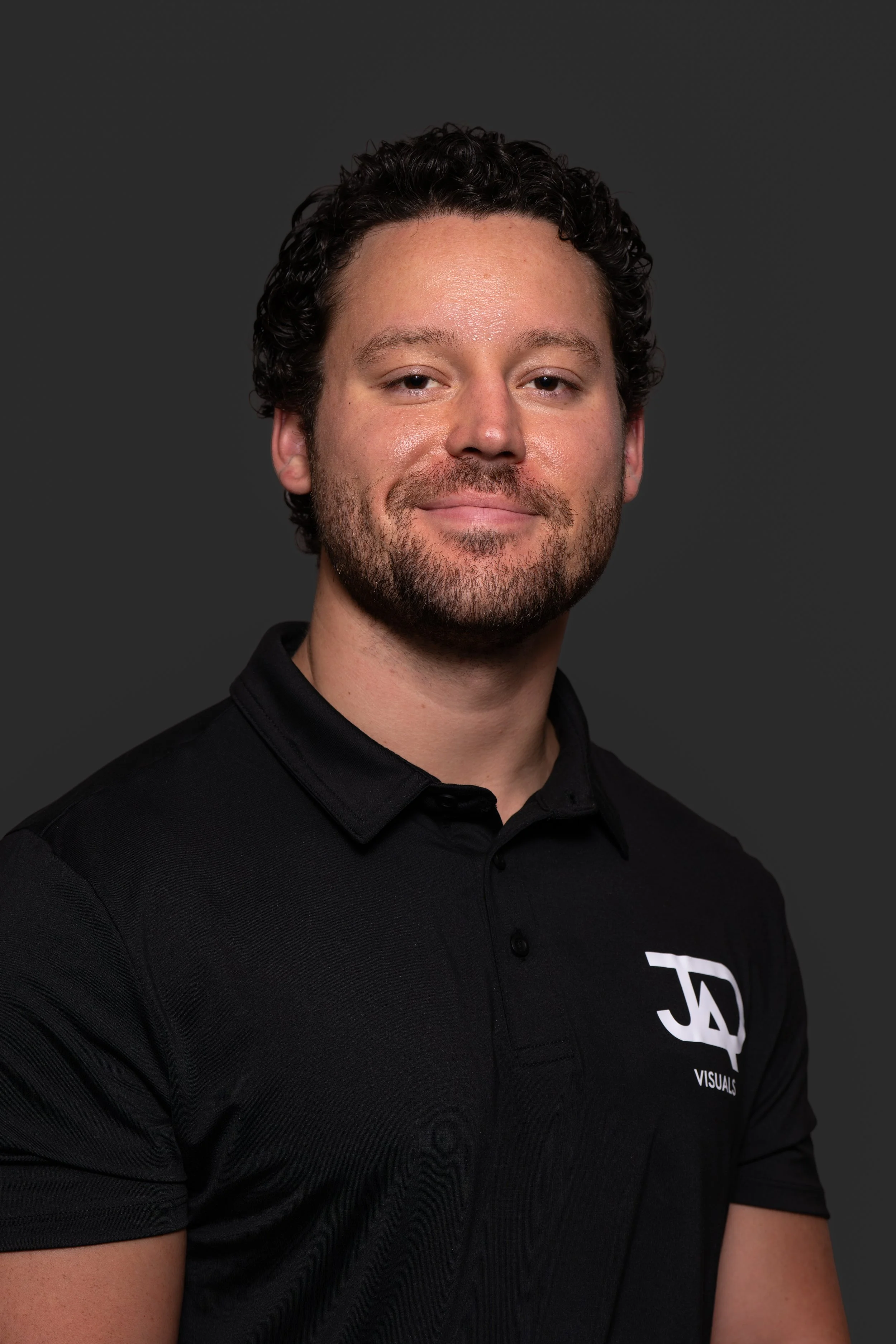 A young man with curly dark hair and a beard, wearing a black collared shirt with a logo on the chest that reads 'JVL Visuals,' posing against a dark gray background.