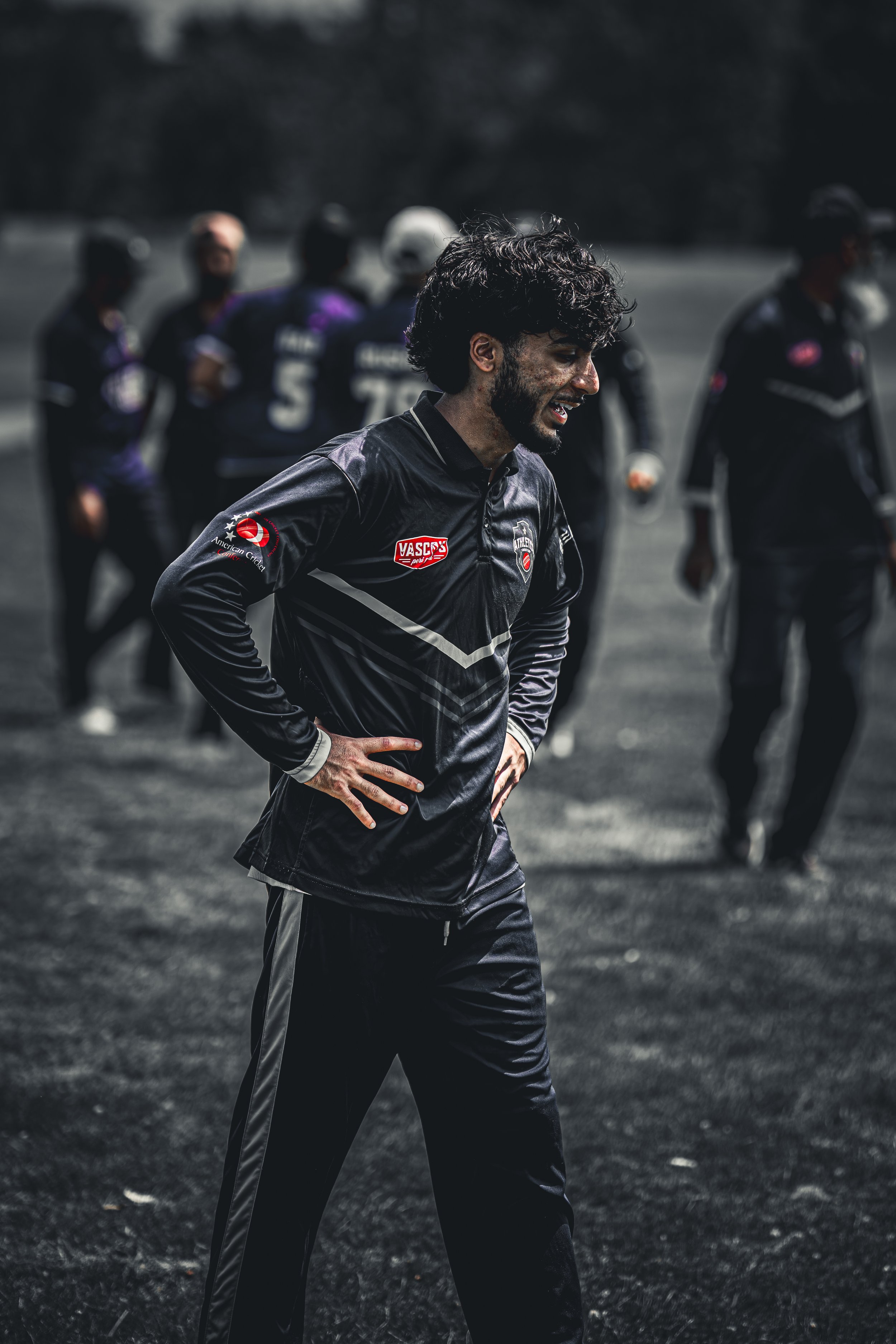 A young man with curly dark hair and a beard, smiling with hands on hips, wearing a black sports jacket with a logo. In the blurred background, other athletes in sports gear are visible, standing outdoors on a dark surface.