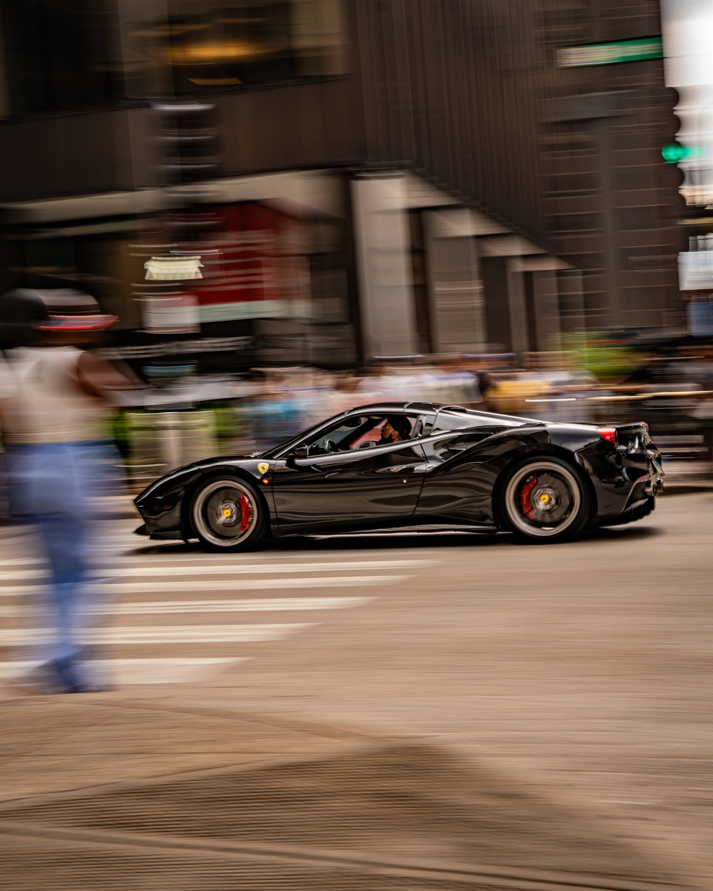 A black sports car with red brake calipers and a low profile driving on a city street with people walking by and blurred buildings in the background.
