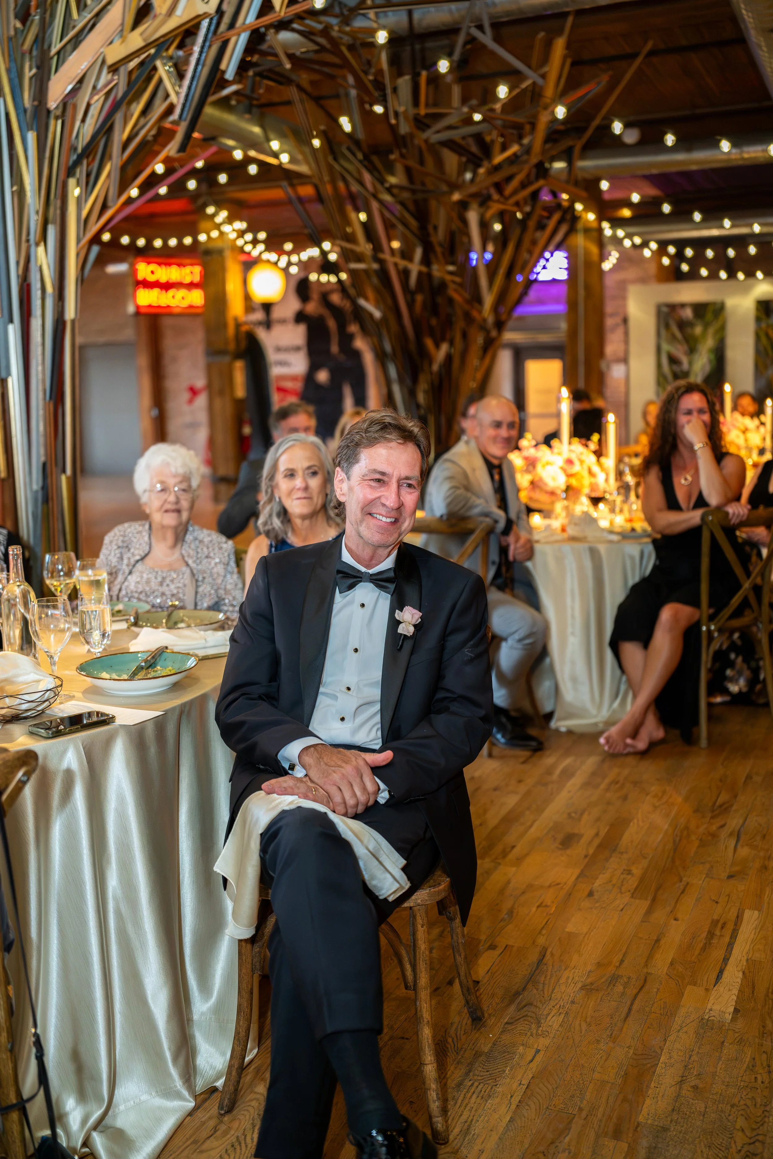 A man dressed in a tuxedo with a bow tie, sitting on a wooden chair at a formal dinner event, smiling with other guests seated at tables decorated with flowers and candles in a warmly lit venue with wooden flooring and brick walls.