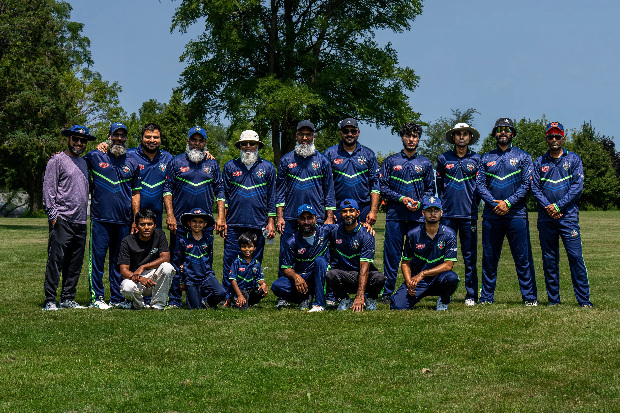 Group of men and boys in matching dark blue sports uniforms with green accents posing outdoors on a grassy area with trees in the background.