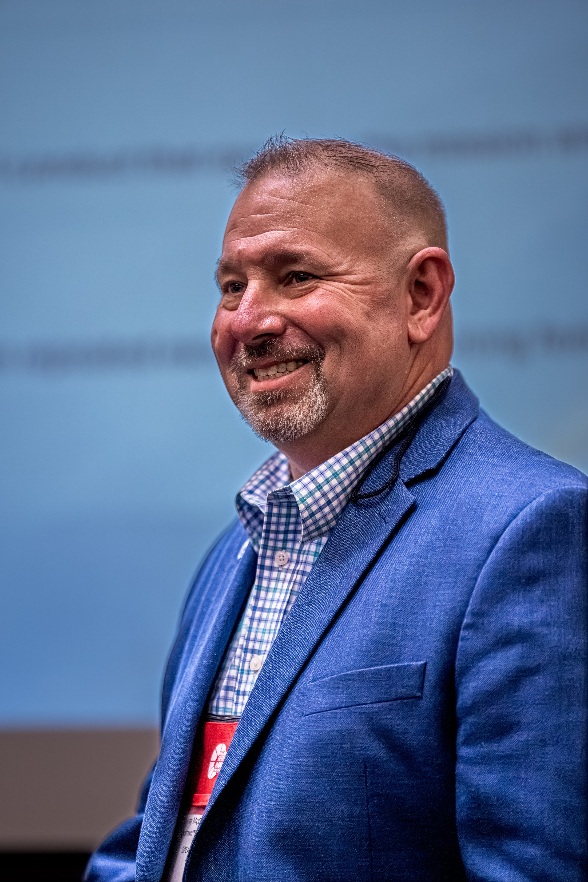 Close-up of a smiling man with short hair, wearing a blue blazer over a checkered shirt, against a blue background.