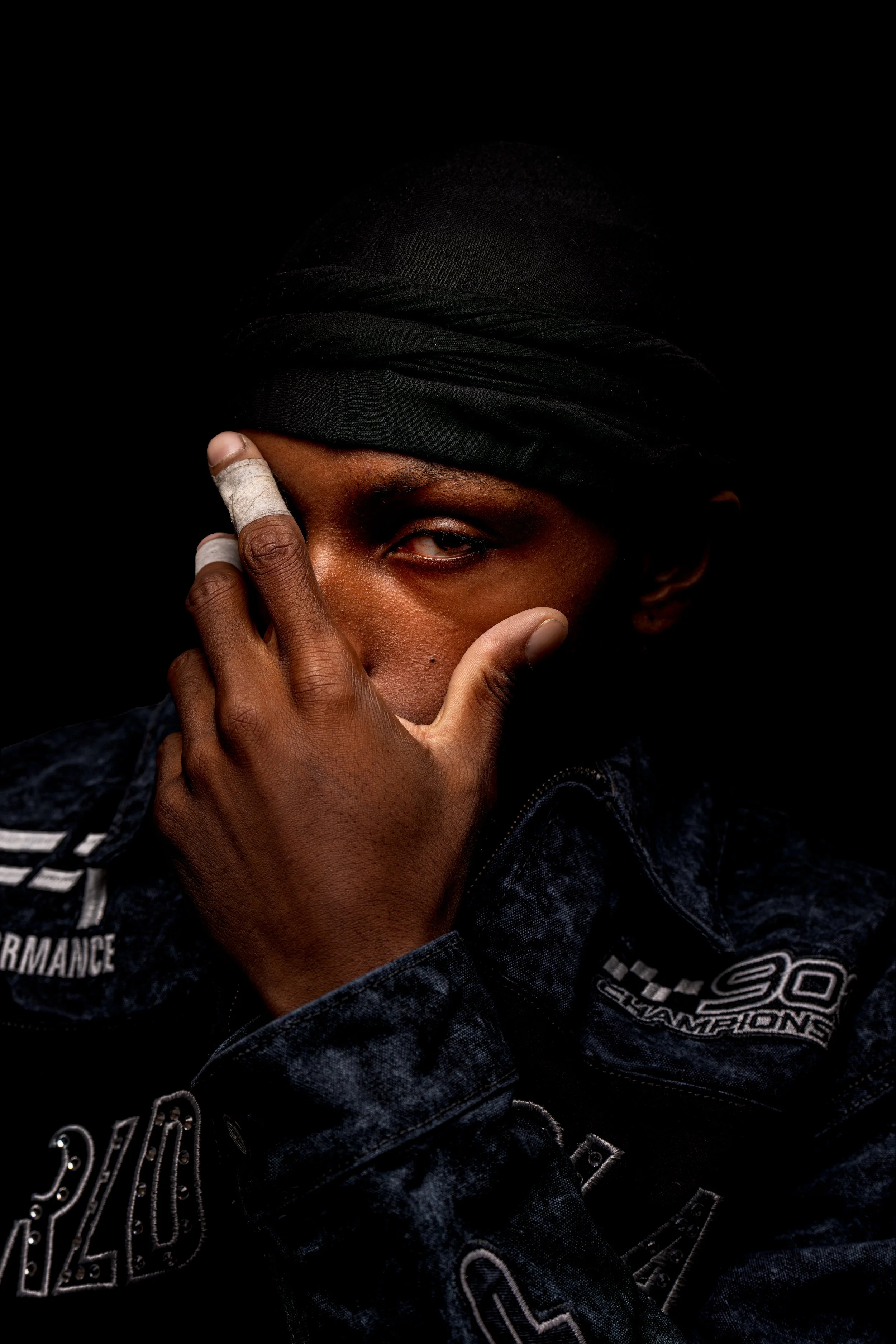 A person wearing a black headscarf, partially covering one eye, and a dark denim jacket with various patches and embroidery, posing against a black background.