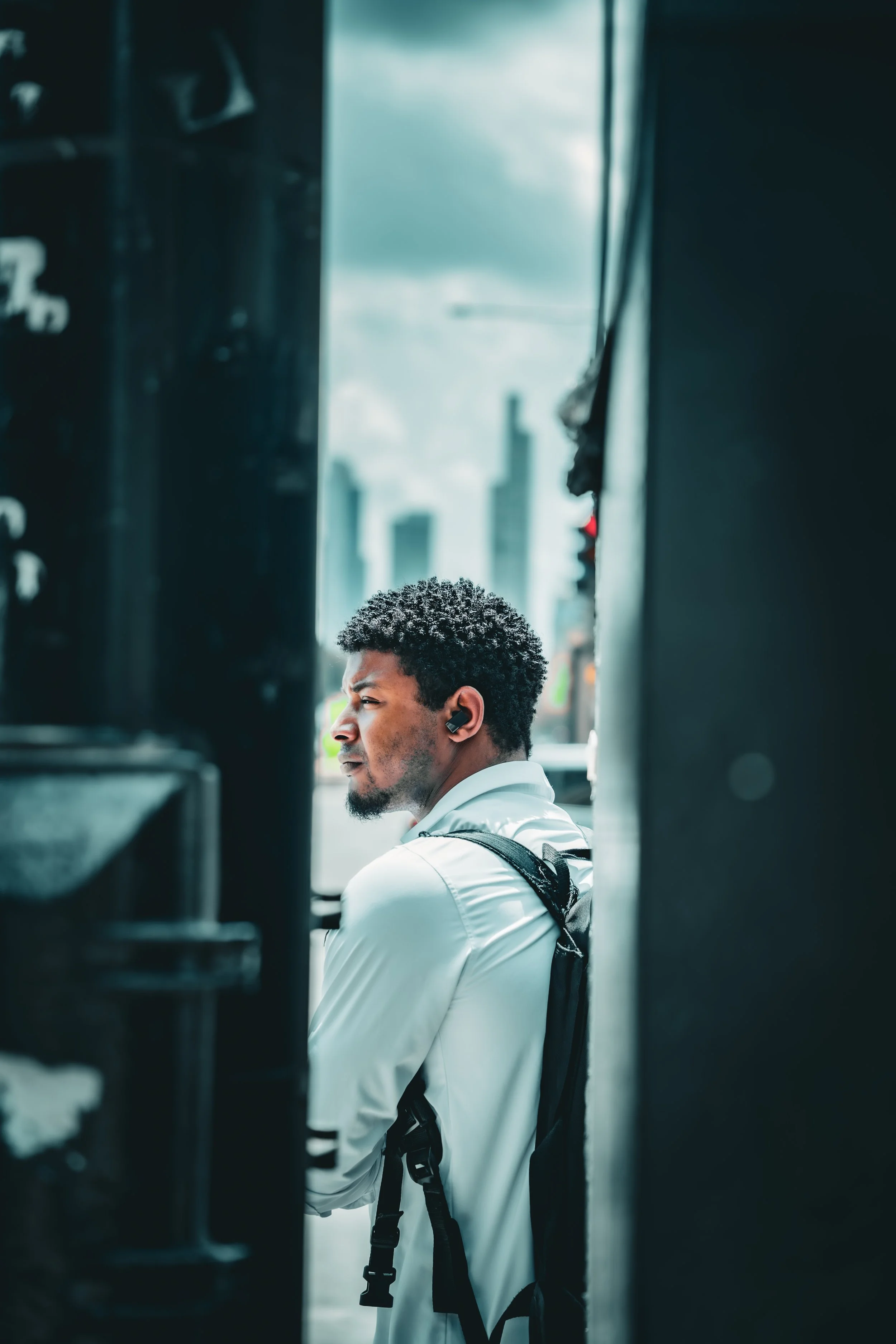 A man with curly black hair and a beard, wearing a white shirt and black backpack, looks to the right through a narrow gap between dark structures outdoors against a cloudy sky.