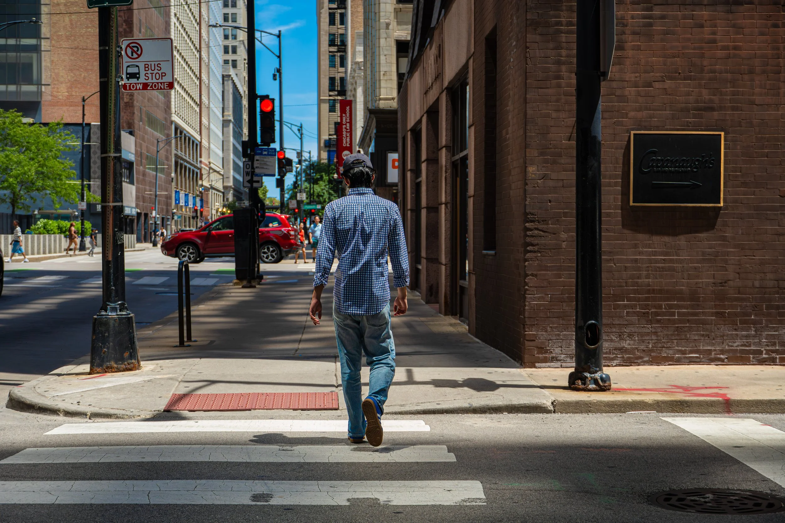 A person with dark hair, wearing a gray cap, a blue and white checkered shirt, and light blue jeans, is walking across a city crosswalk on a sunny day. The background features tall buildings, a red car, and a street with other pedestrians. There are 