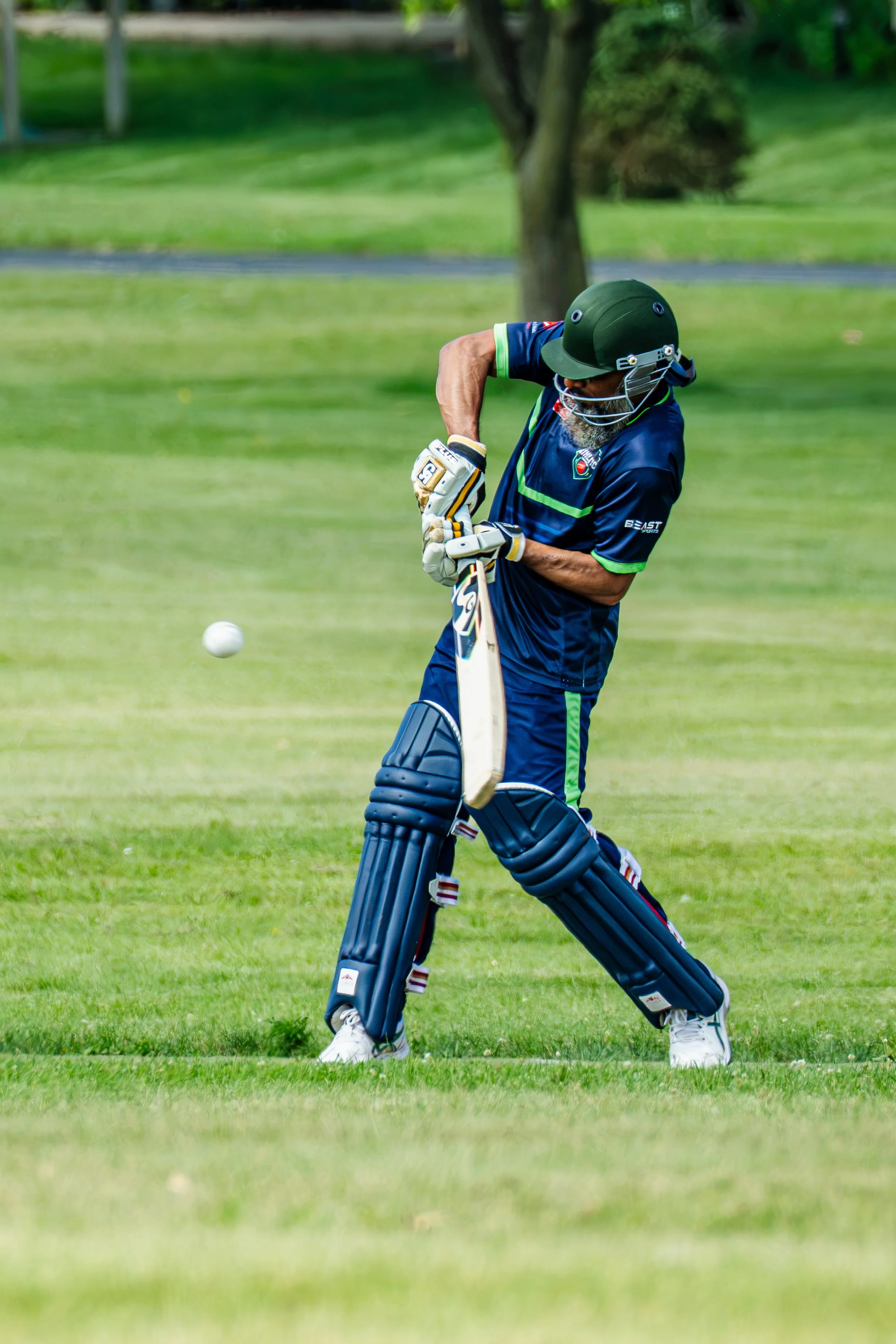 A man playing cricket on a grassy field, wearing protective gear including pads, gloves, and a helmet. He is in a batting stance with a cricket bat, focusing on the approaching ball.