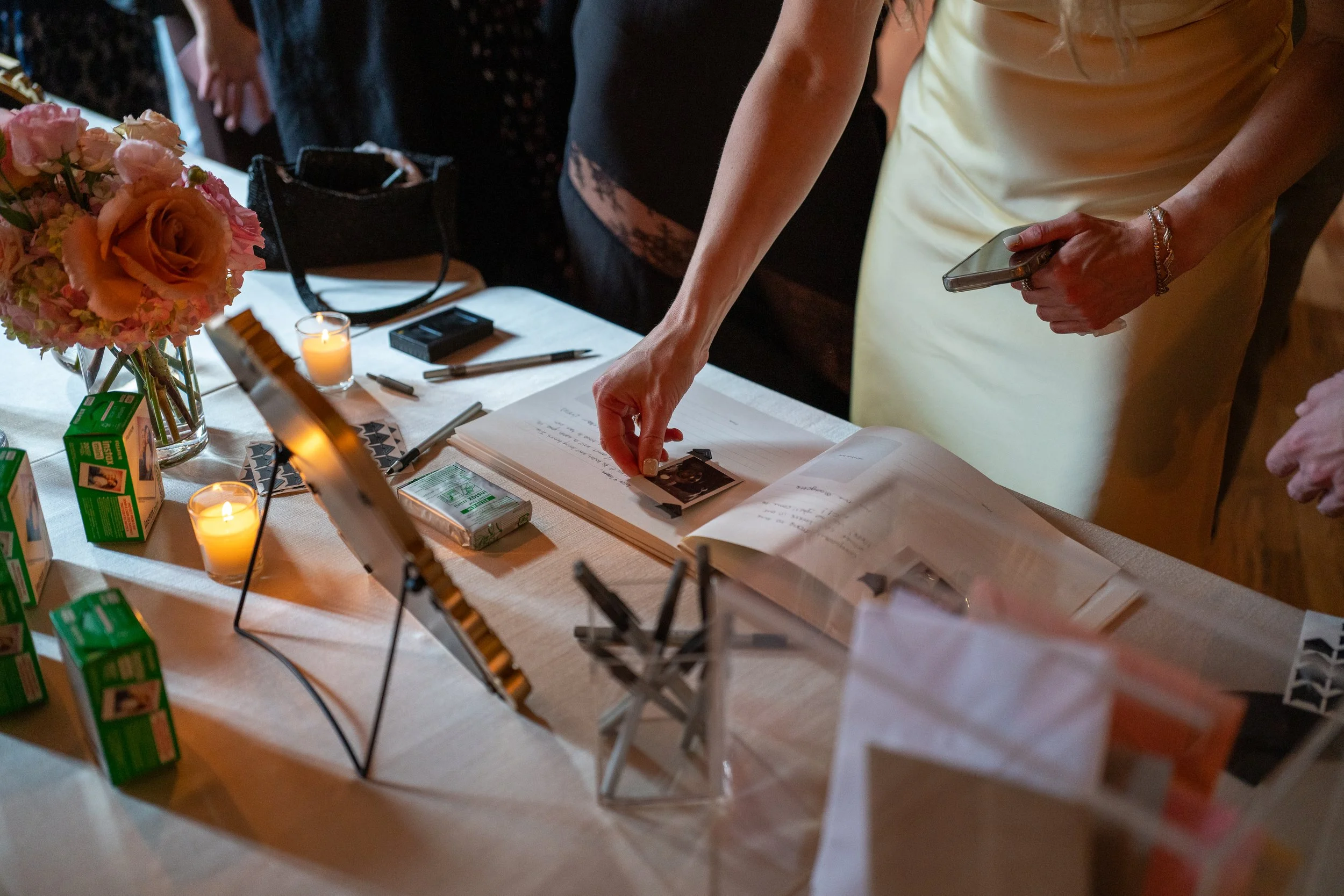 A woman in a yellow dress reaching for a photo on a table with flowers, candles, markers, and small boxes or cartons in a dimly lit setting.