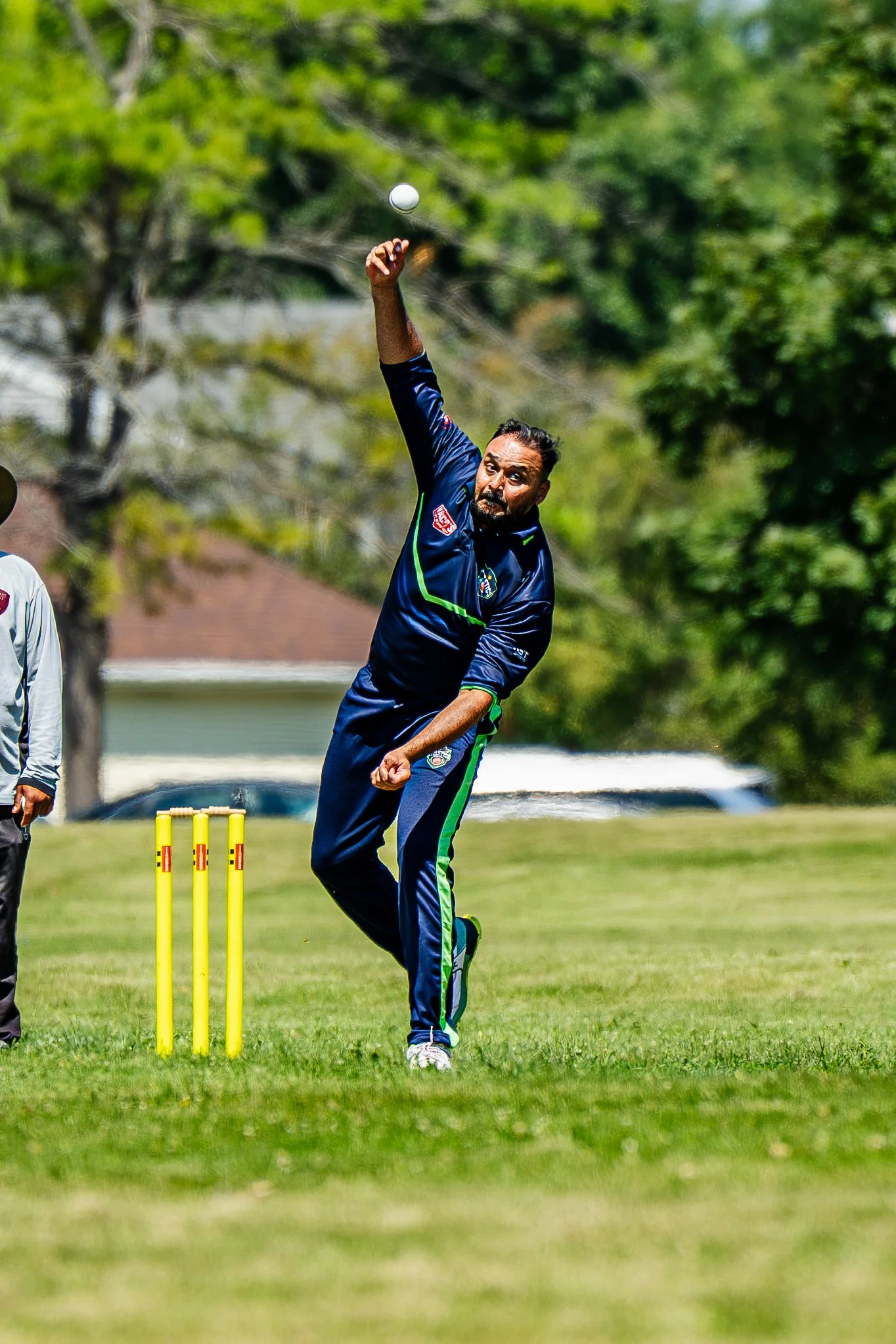 A man in a sports uniform is bowling a cricket ball during a game on a grassy field. He appears focused, with trees in the background.