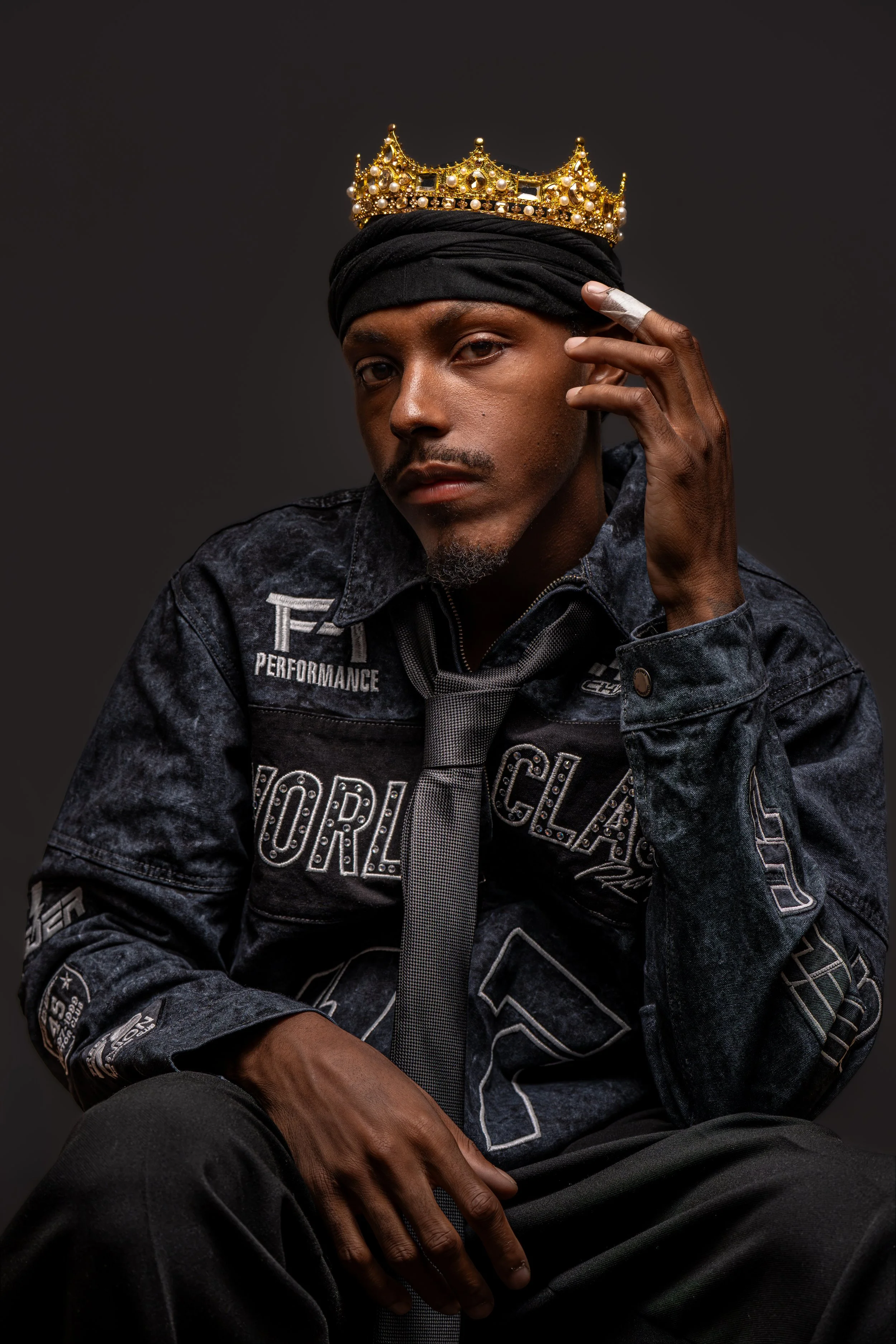 A young man with a dark complexion wearing a black headwrap, dark denim jacket with patches, and a gray tie. He is touching his face near his temple, and he has a serious expression. A gold crown adorned with pearls and small gemstones is placed on h