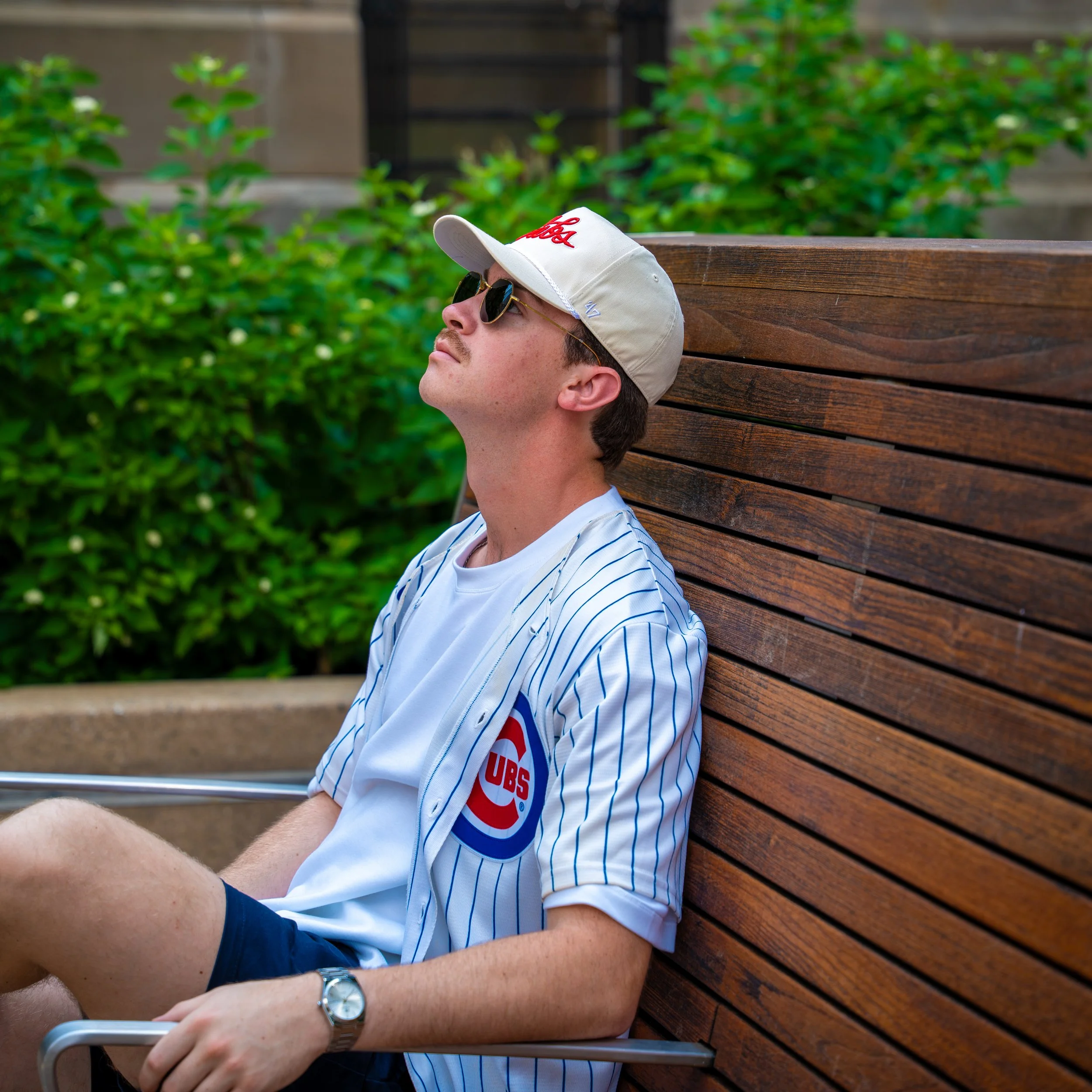 A young man with a beard, wearing sunglasses and a white baseball cap with red lettering, sitting on a wooden bench outdoors, looking upward.
