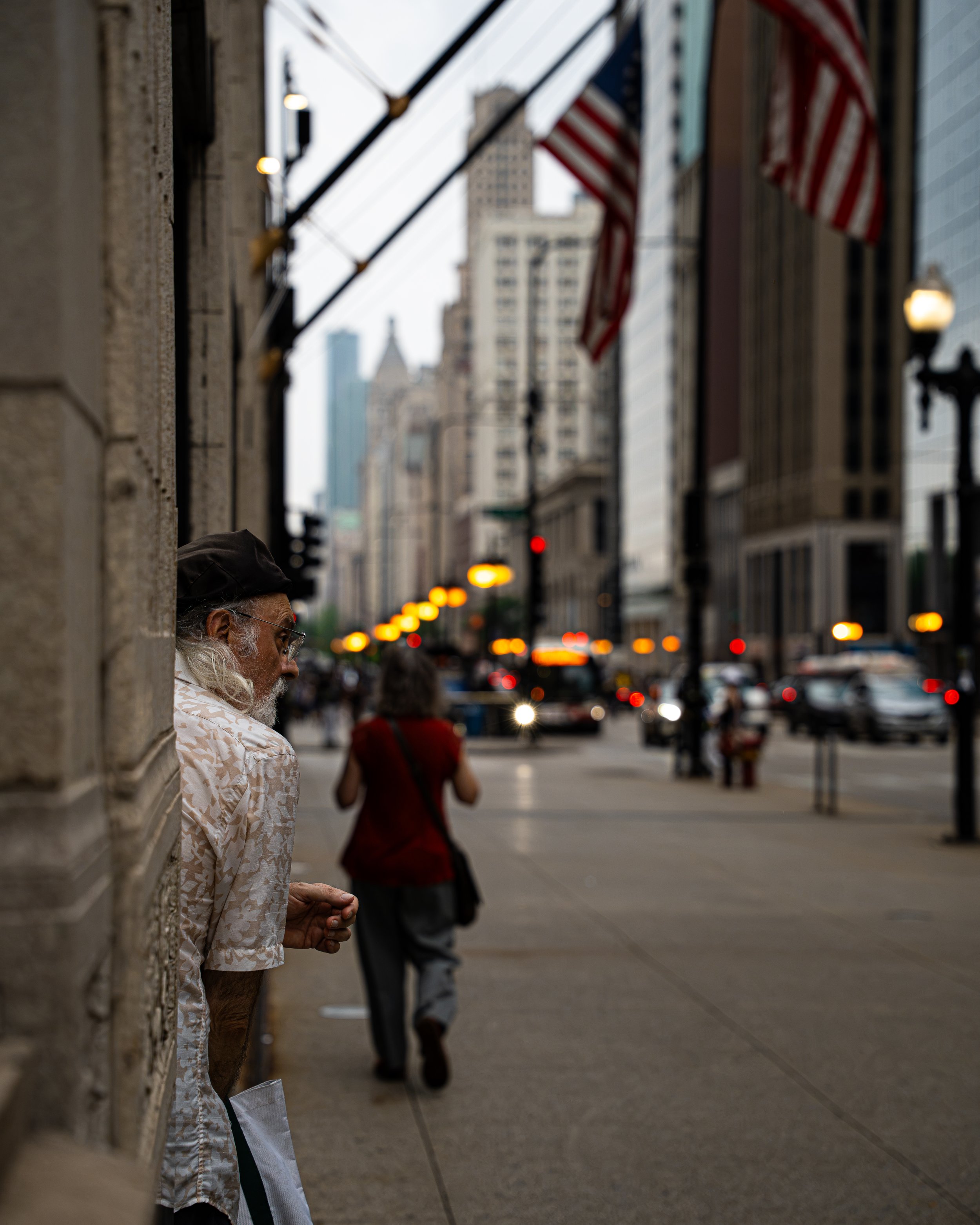 A man with long hair and glasses stands against a textured building wall on a city street, with various people and tall buildings in the background, some US flags hanging from the building.