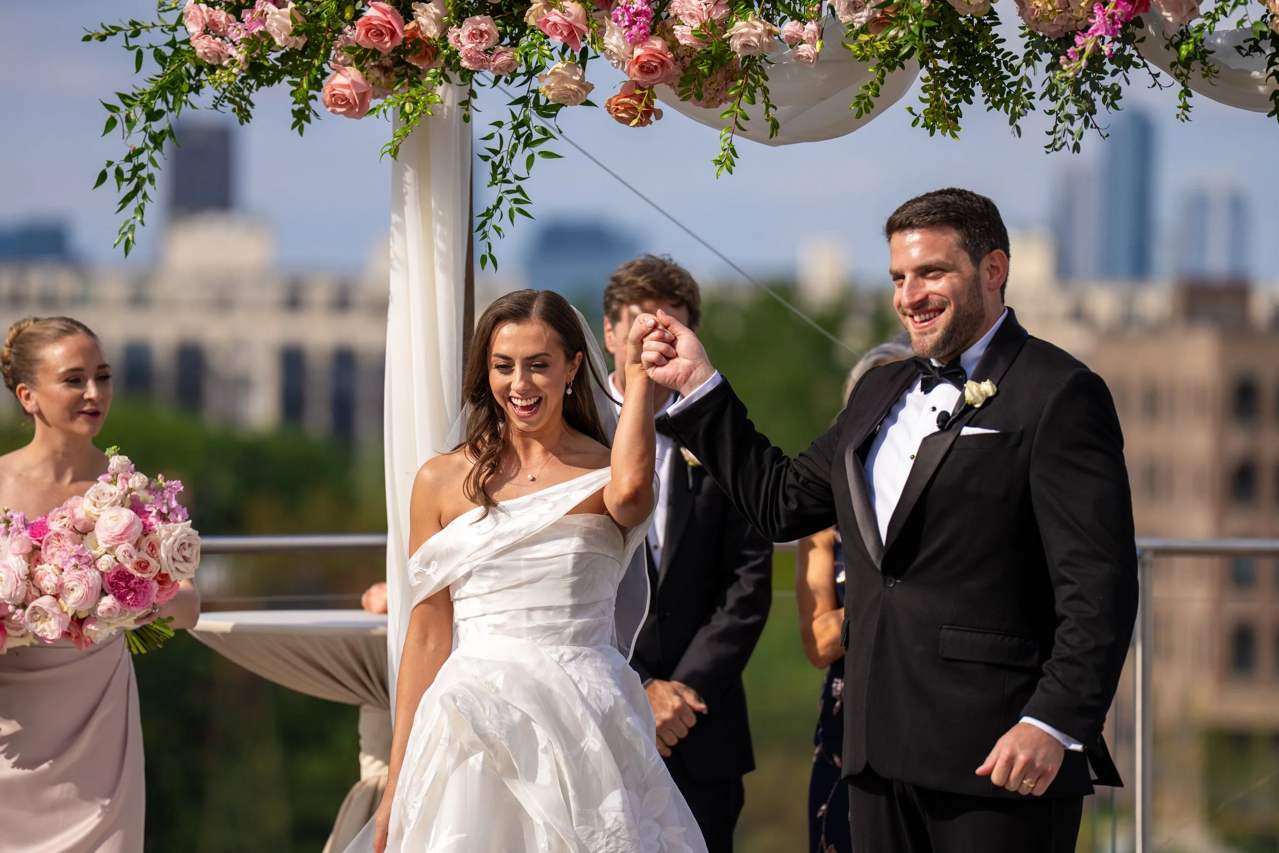 A bride and groom holding hands and smiling at a wedding ceremony outdoors, with a floral decoration overhead and city skyline in the background.