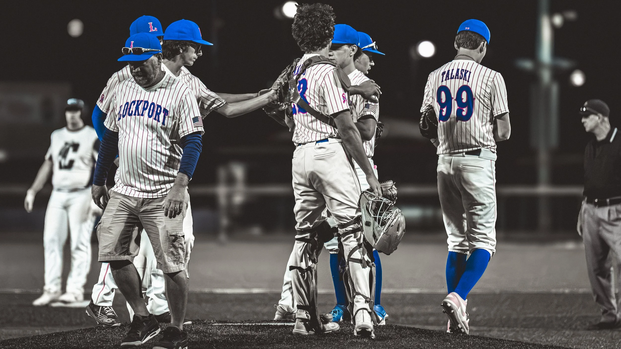 A group of baseball players in striped uniforms and blue caps gathering on the field at night, with one player wearing catcher's gear.