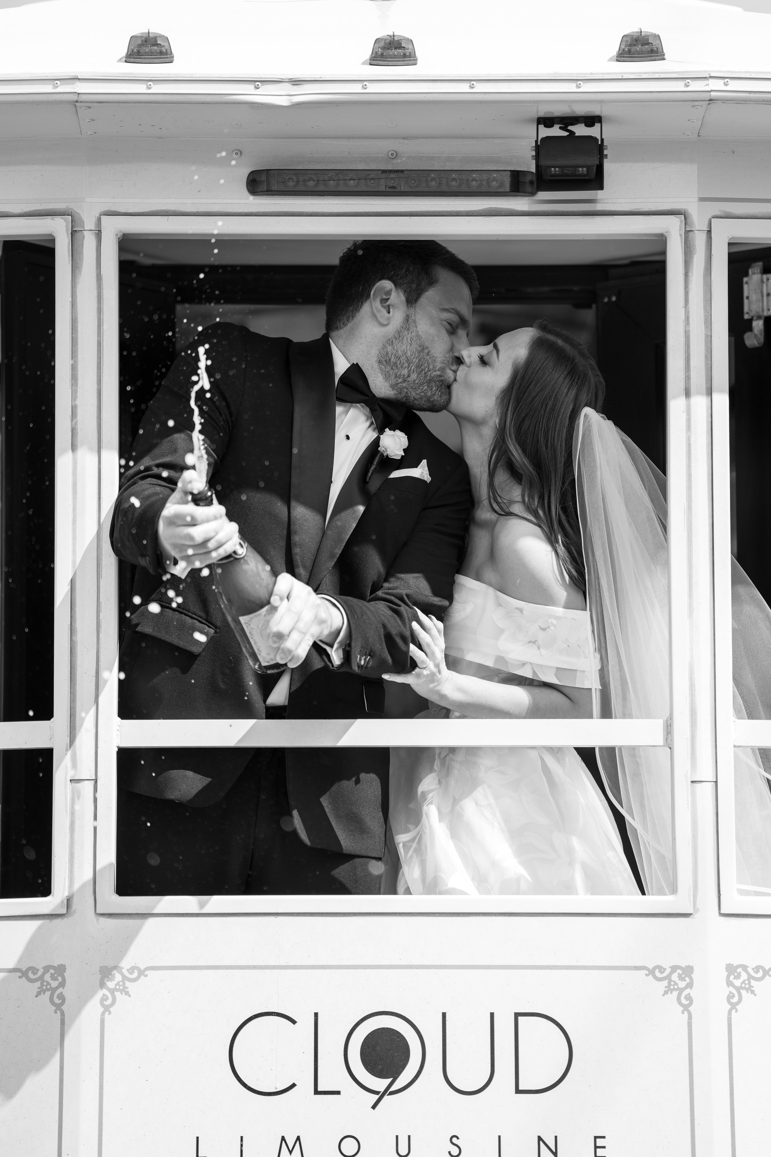 A black-and-white photo of a couple in wedding attire sharing a kiss inside a horse-drawn carriage, the groom holding a bottle with champagne spray.