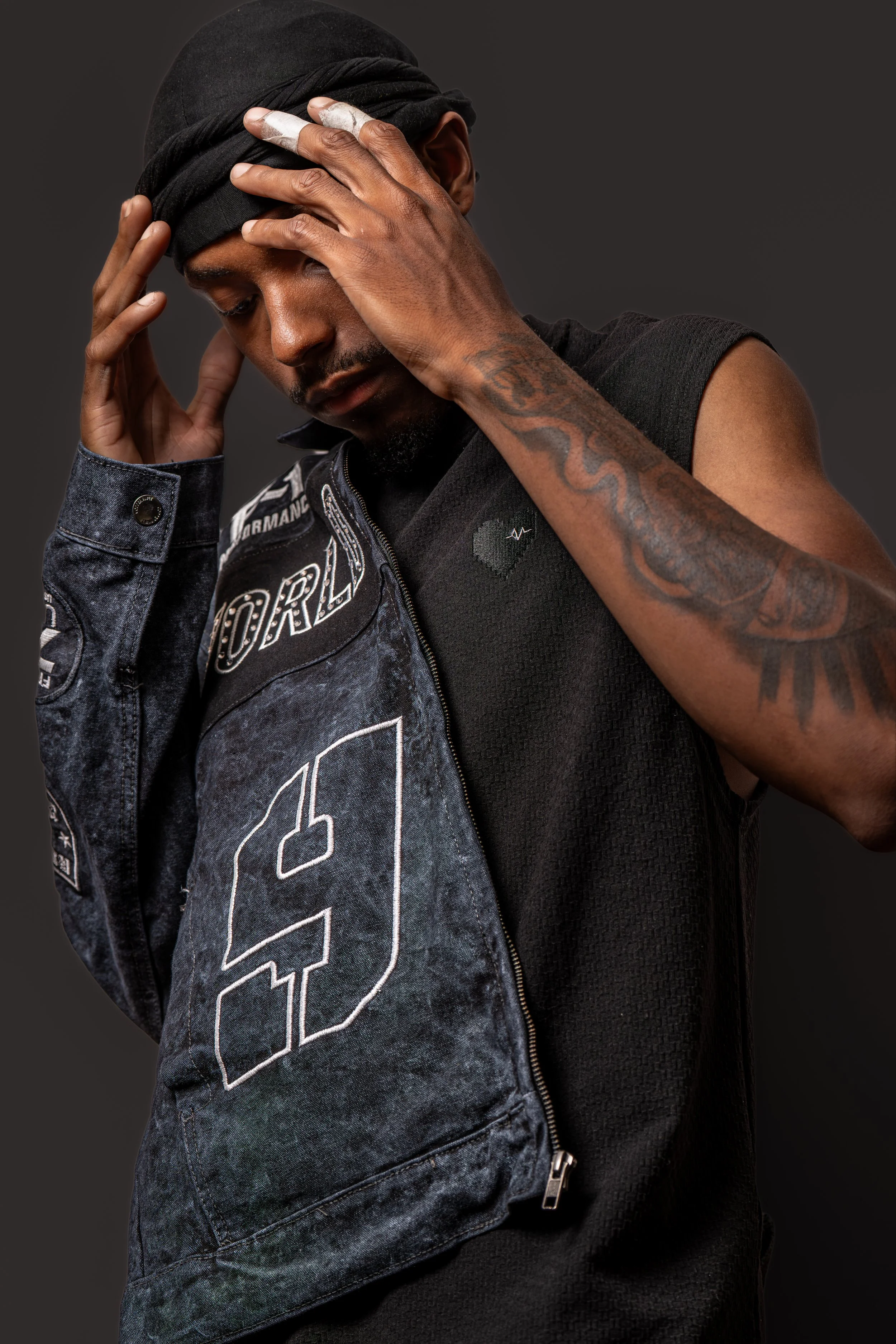 A man with a dark head wrap and tattoos on his arms, wearing a sleeveless black shirt and a denim vest with patches, holding his head with both hands and looking down, with a serious expression against a dark gray background.