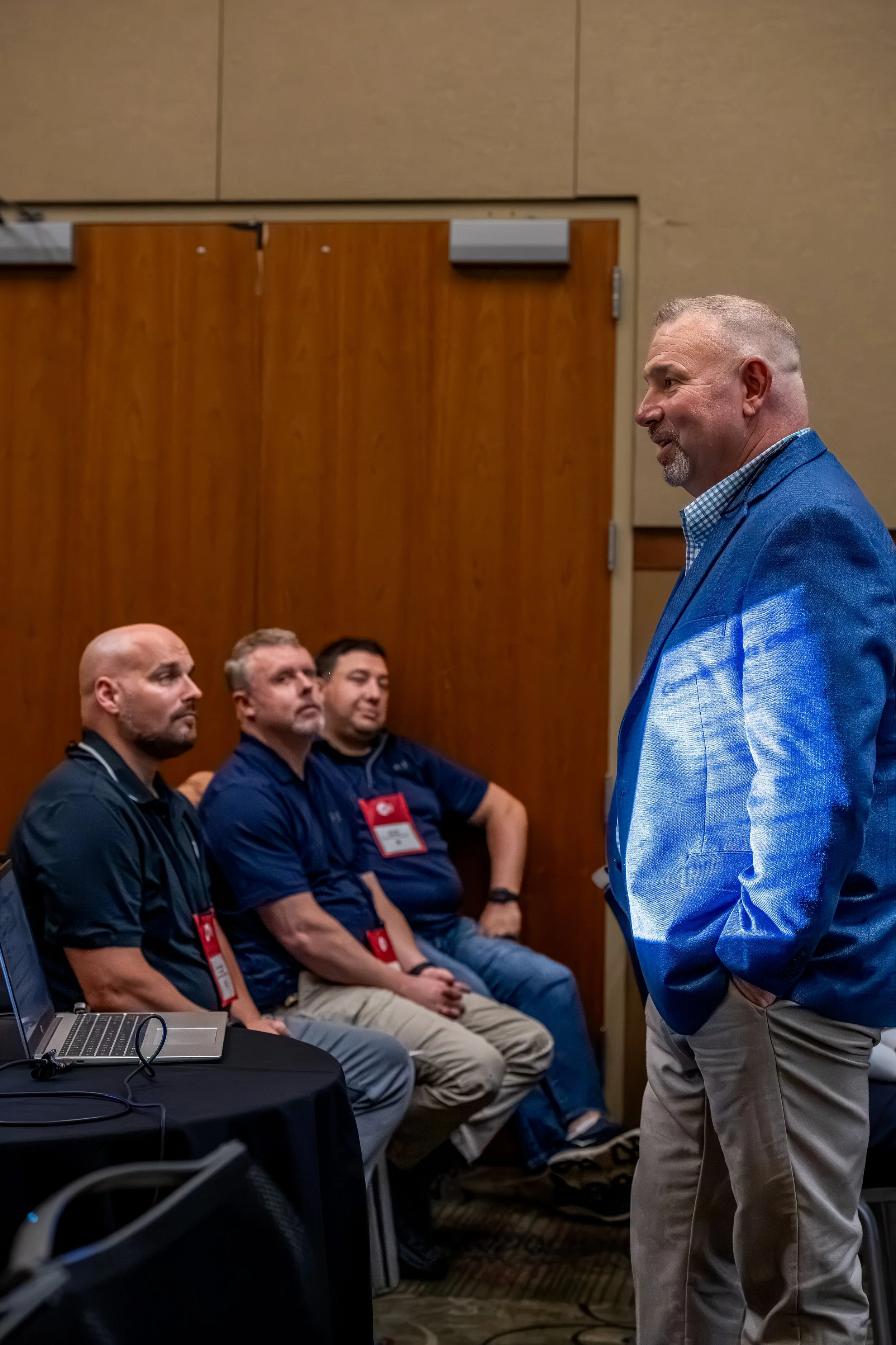 A man in a blue blazer and beige pants is standing and speaking, while four men sit and listen attentively at a conference or seminar. There is a laptop and some cables on a nearby table.