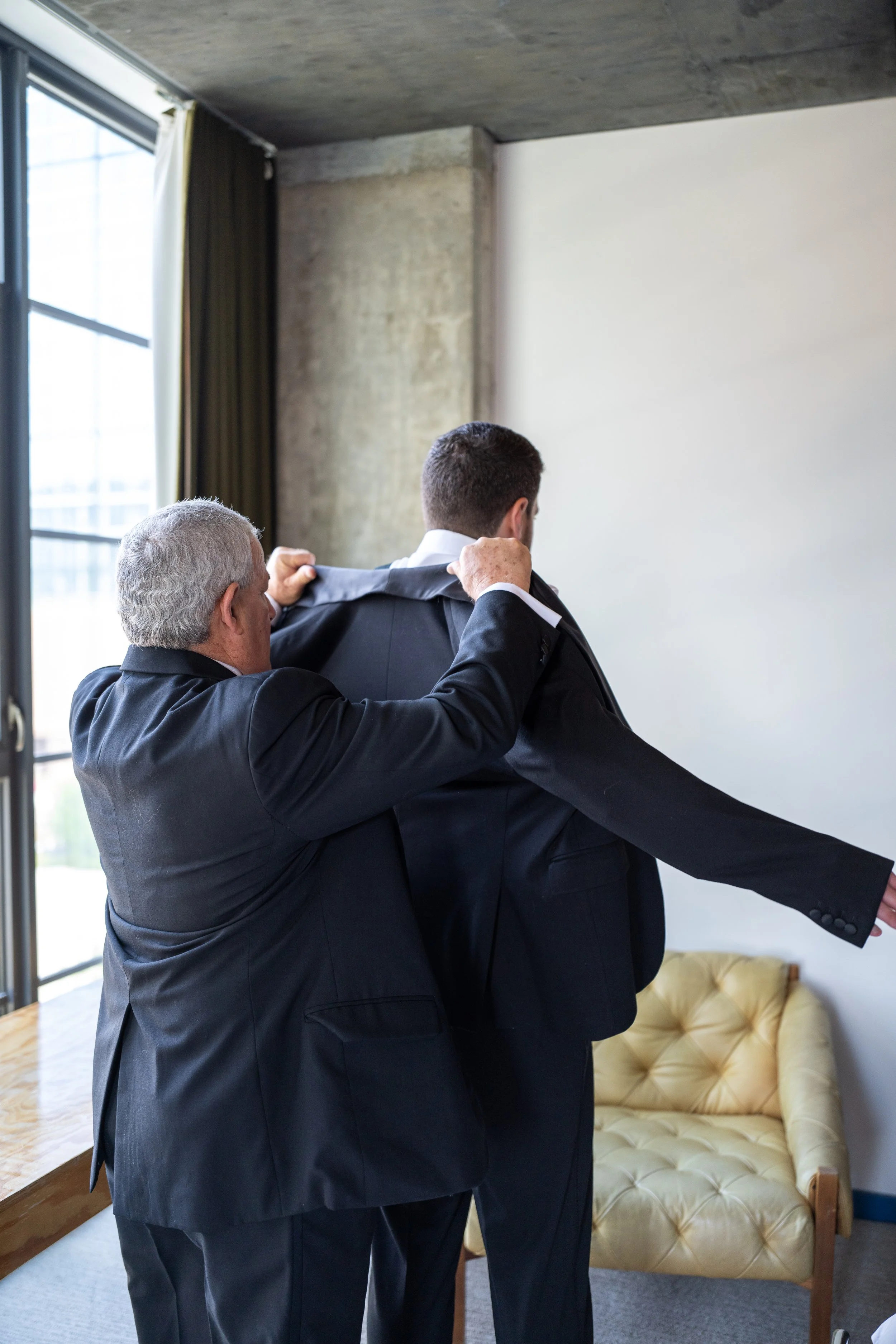 An older man helping a younger man put on a suit jacket, standing in a room with natural light, a large window with curtains, and a yellow tufted sofa.