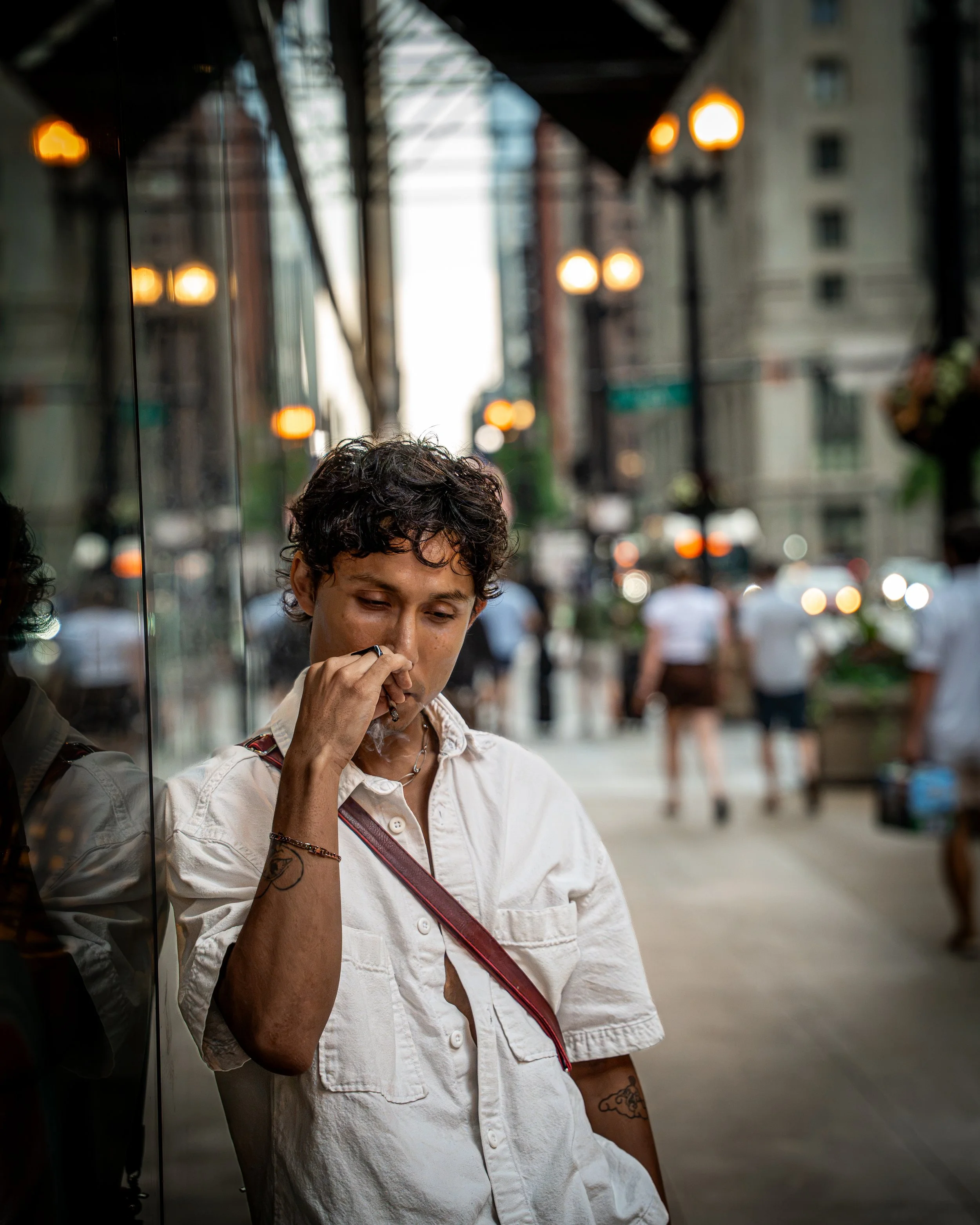 A young woman with short curly hair, wearing a white button-up shirt, smoking a cigarette on an urban street, with blurred city buildings, streetlights, and pedestrians in the background during daytime.