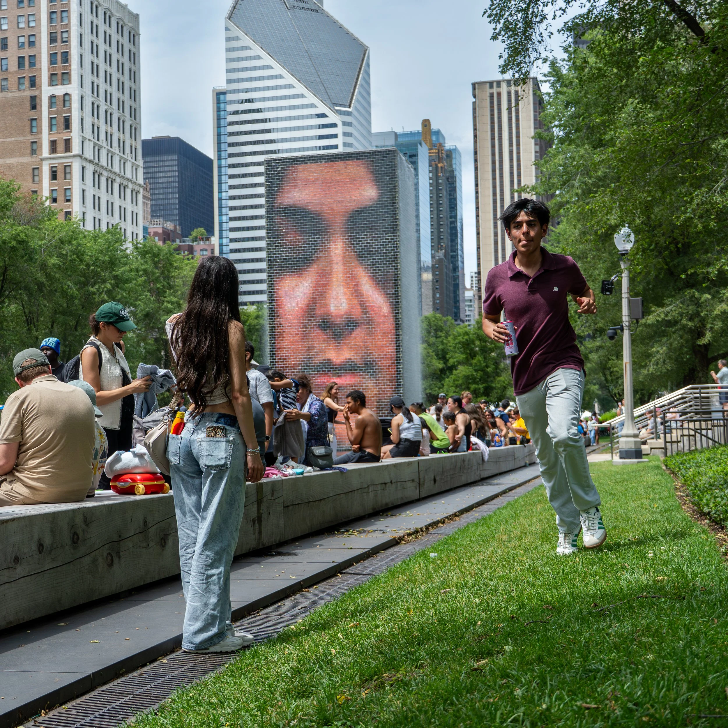 People gathered in a park with tall buildings in the background, including a large pixel art mural of a woman's face on a building. Some individuals are sitting, while others are walking or standing, and a man in a maroon shirt is holding a bottle of