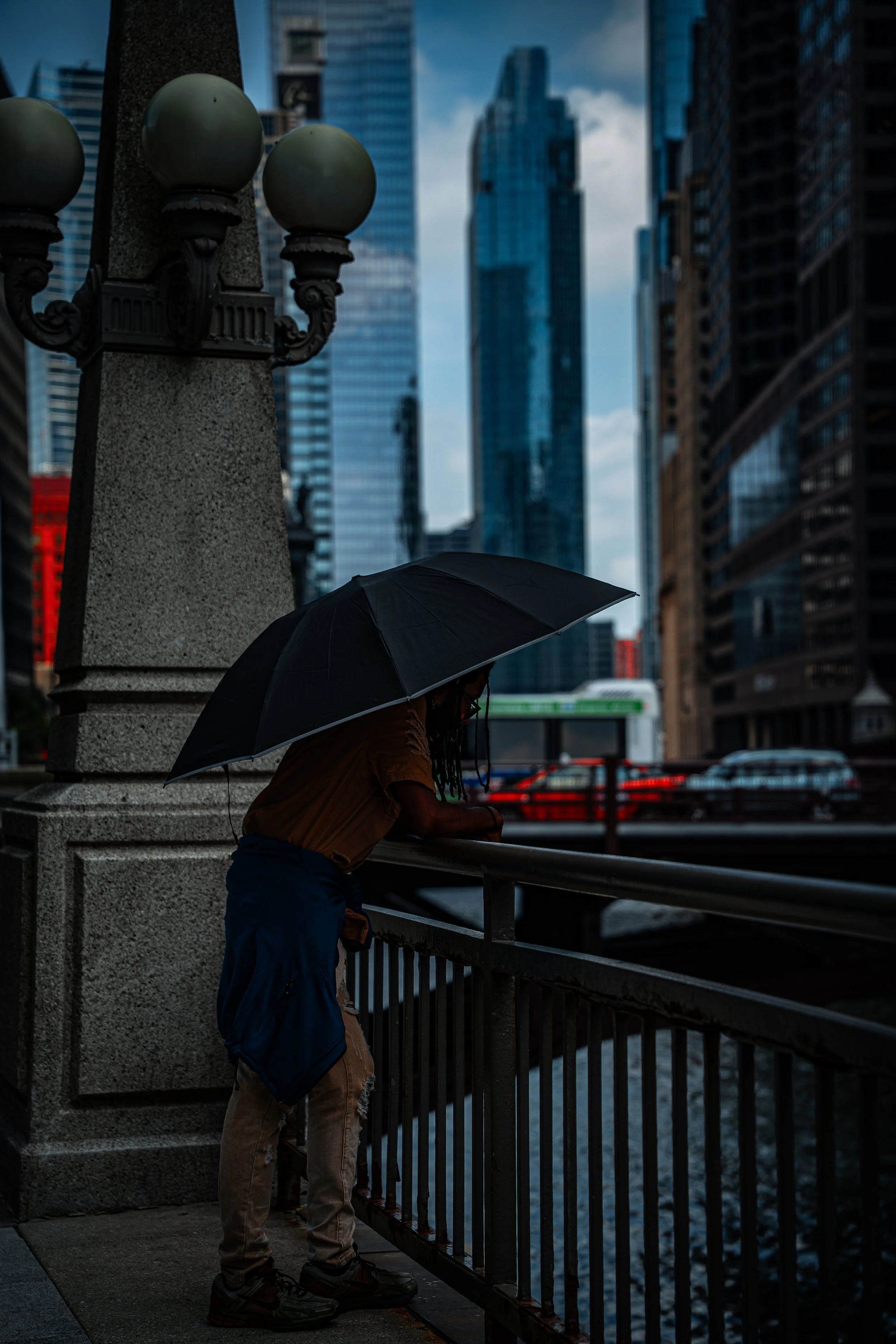 A person with a black umbrella standing on an urban bridge, with tall skyscrapers in the background and cloudy sky.