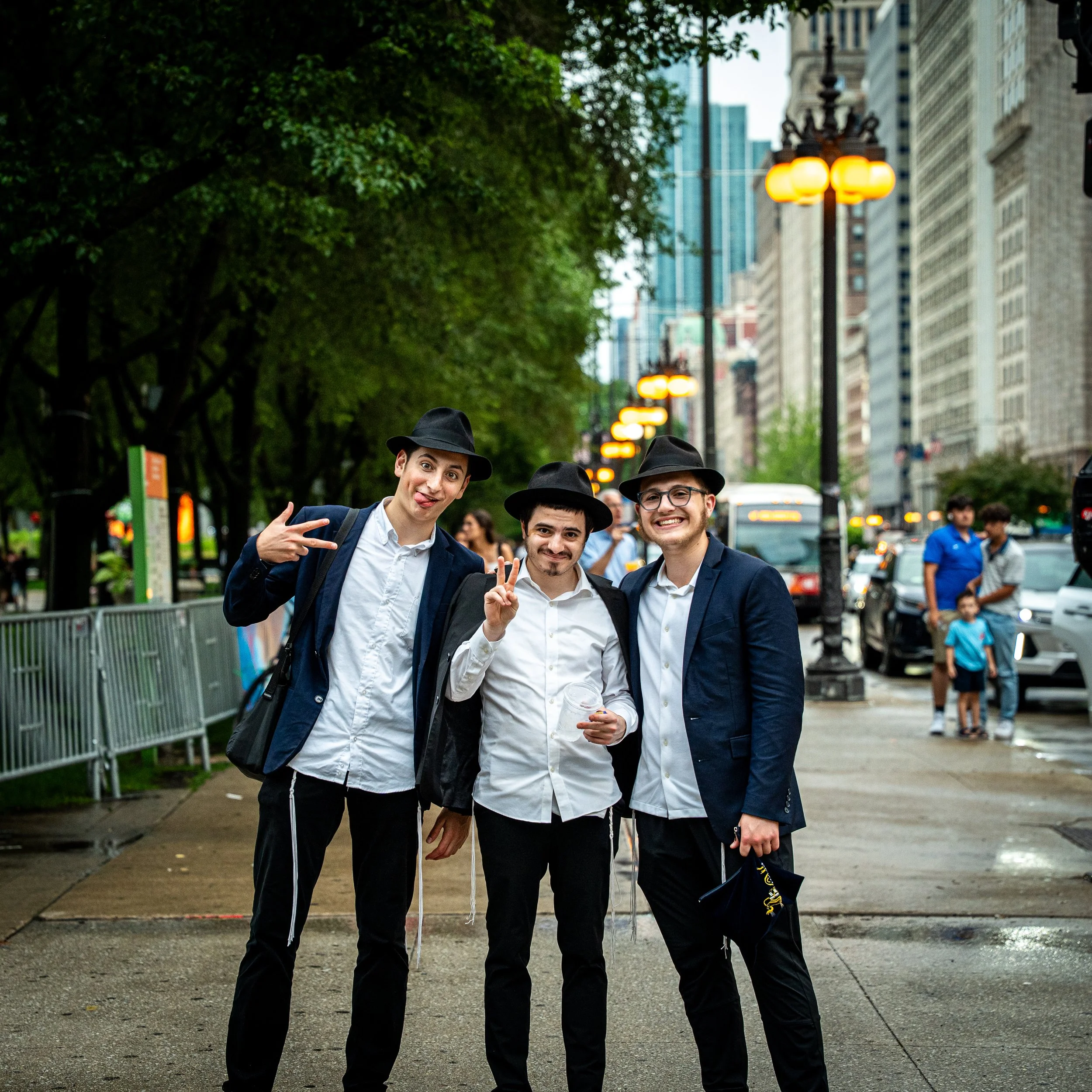 Three young men dressed in suits and white shirts, wearing black hats, standing together outdoors in a city street, smiling and posing for the photo with city buildings in the background.