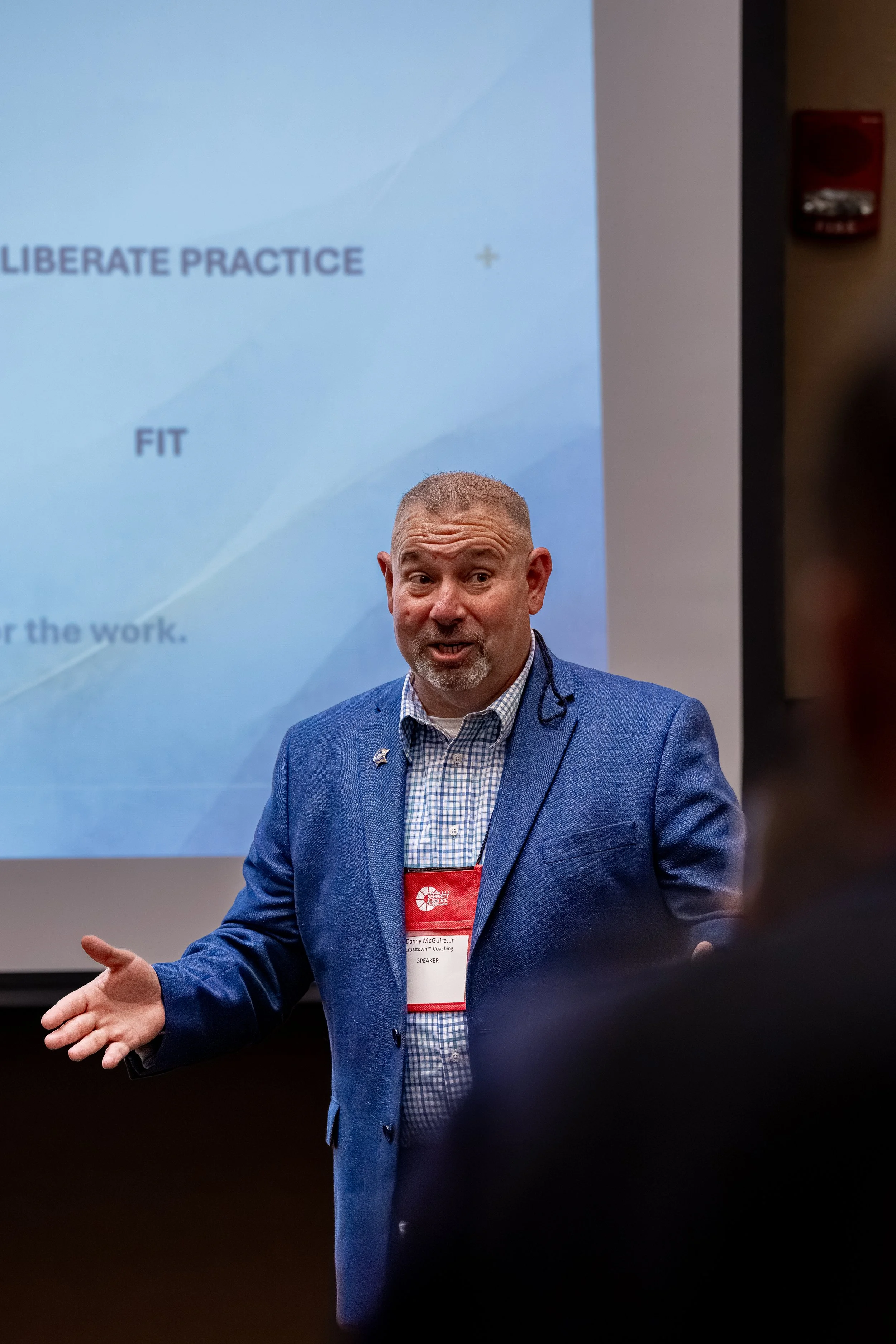 A man in a blue blazer giving a presentation in front of a blue slide with the words 'LIBERATE PRACTICE' visible. The man is gesturing with his left hand and has a conference badge hanging around his neck.