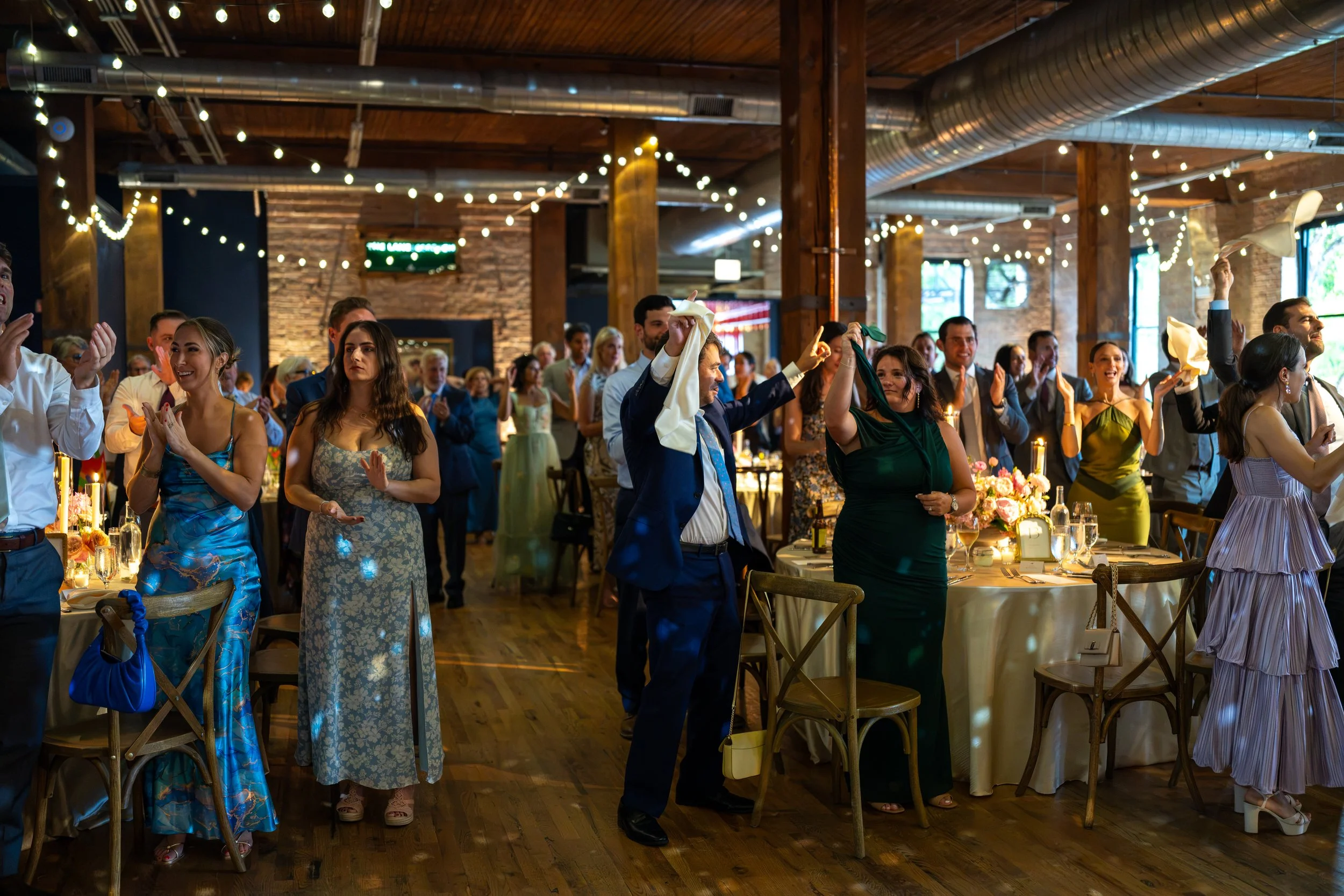 People dancing and enjoying themselves at a reception or party in a rustic venue with string lights and decorated tables.