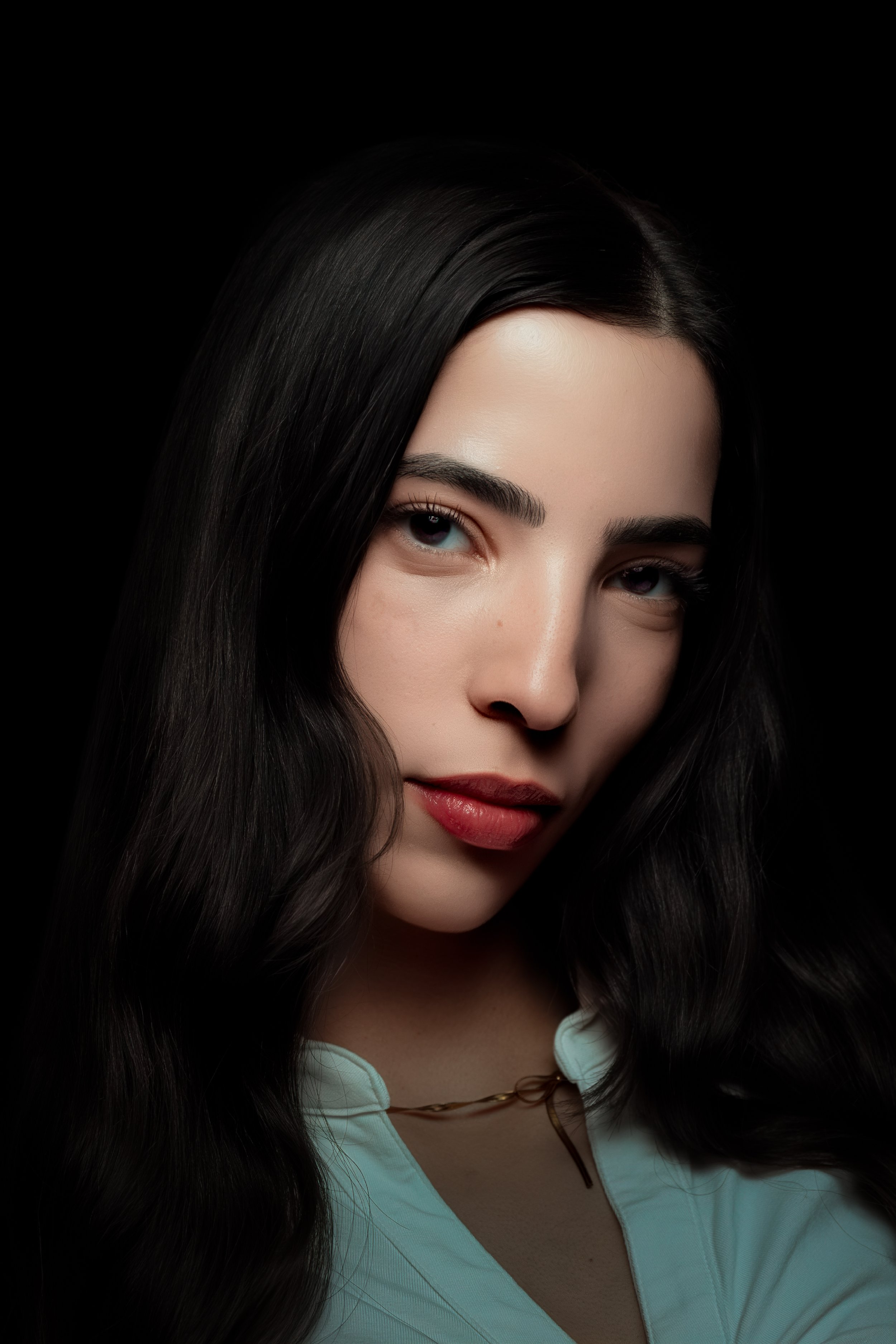 Close-up of a woman with long, dark hair, light skin, and makeup, wearing a white top and a jewelry chain, against a black background.