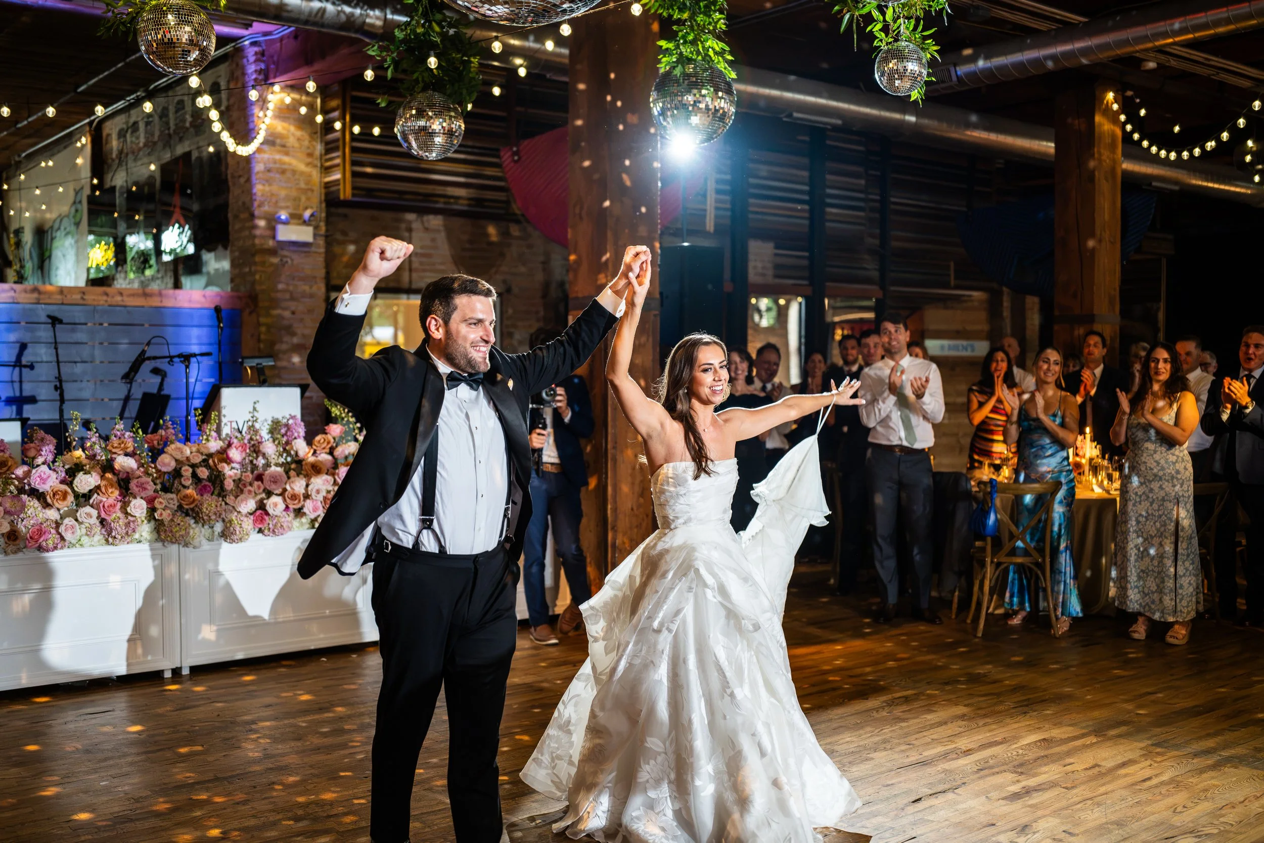 A newlywed couple dancing at their wedding reception, surrounded by friends and family. The groom is wearing a black tuxedo and the bride a white wedding gown, smiling and celebrating in a decorated, festive venue with disco balls and string lights.