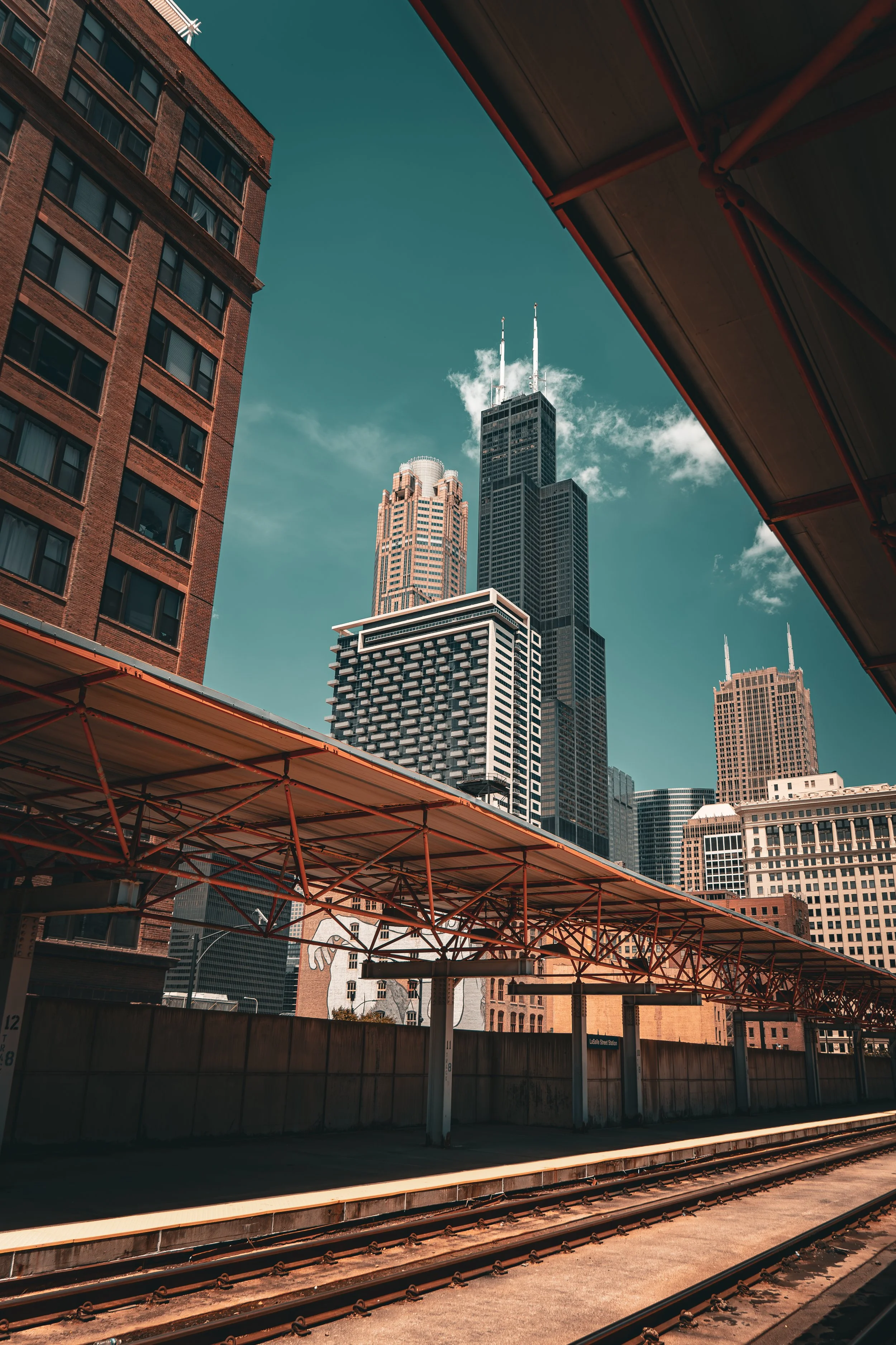 View of skyscrapers in a city skyline with the train station platform and tracks in the foreground, and a blue sky with some clouds.