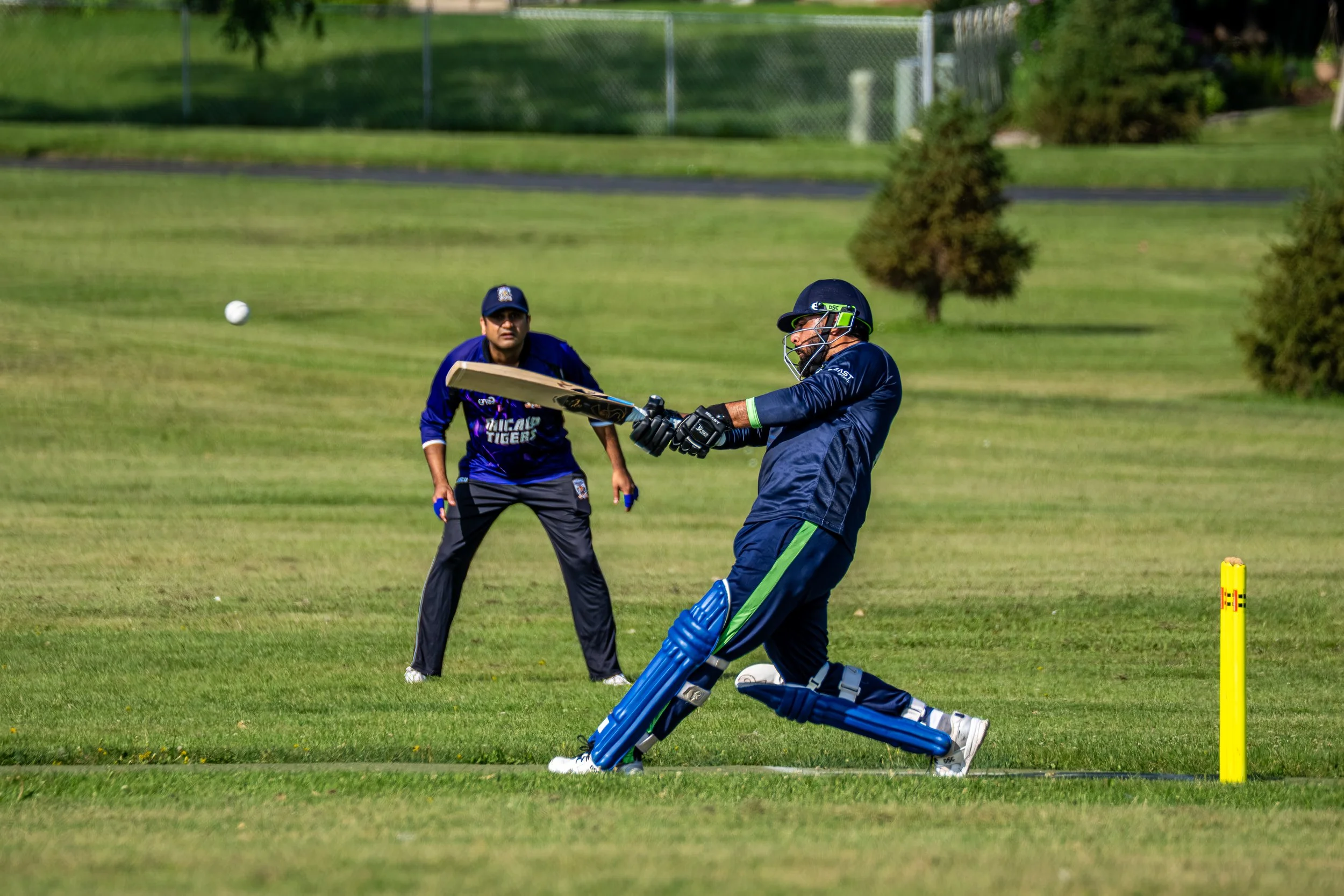 A cricket batter is in front of the wicket, preparing to hit the ball, while a wicketkeeper crouches behind the wicket in a protective stance during a game on a grassy field.