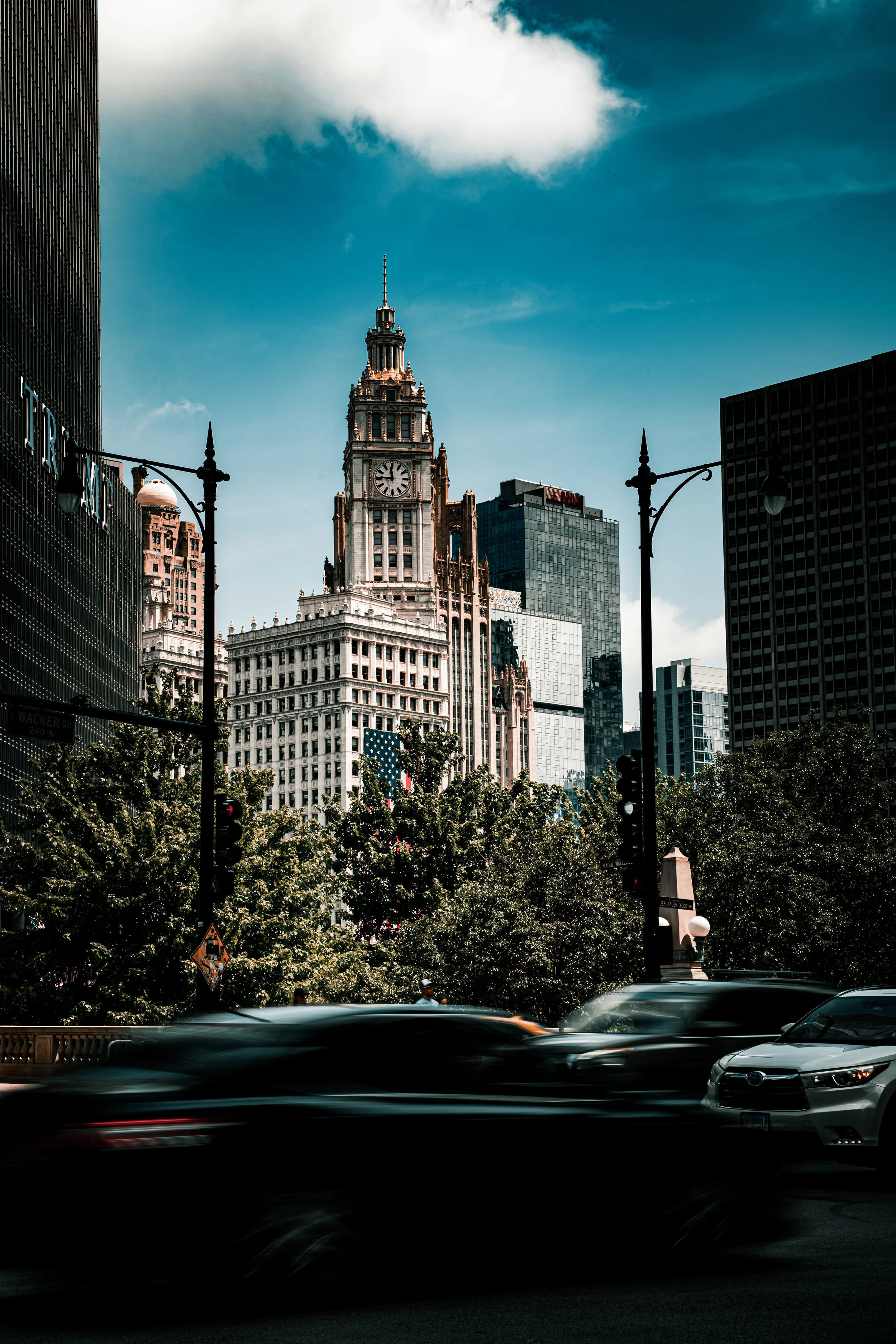 City street scene with tall historic clock tower building, modern skyscrapers, trees, streetlights, moving cars, and a cloudy sky.