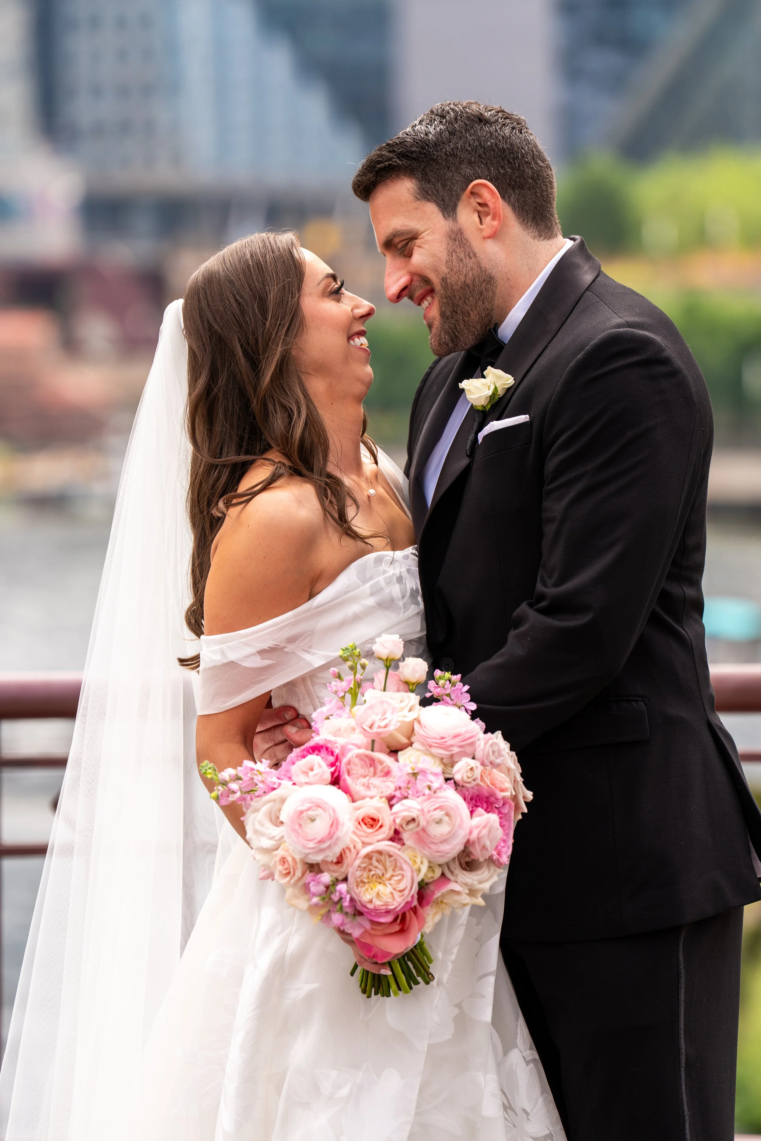 A happy bride and groom standing close together, smiling and looking into each other's eyes, with the bride holding a large bouquet of pink and white flowers, outdoors in a city setting.