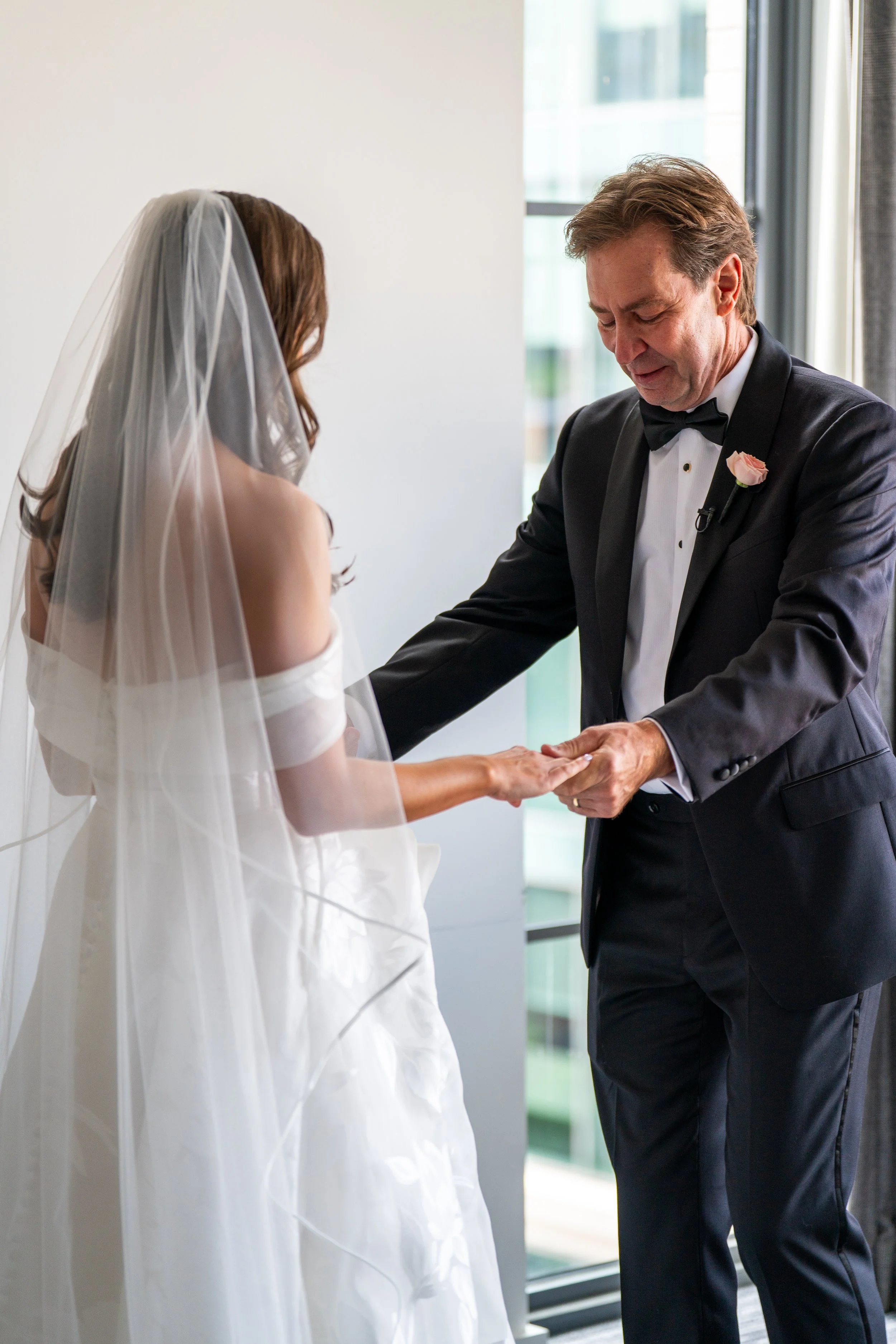 A groom in a black tuxedo with a pink boutonniere is placing a wedding ring on a bride's finger. The bride is wearing a white wedding dress and veil, standing with her back to the camera. The scene takes place indoors near a large window with natural