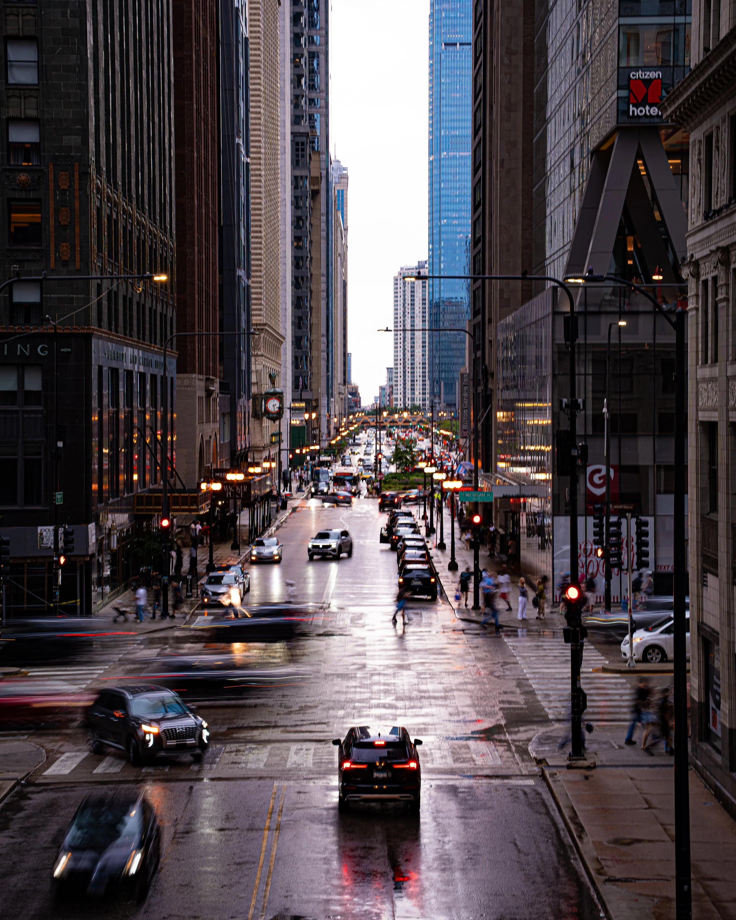 A city street scene in downtown during dusk or early evening, with tall buildings on both sides, cars and pedestrians, and streetlights illuminating the wet pavement.