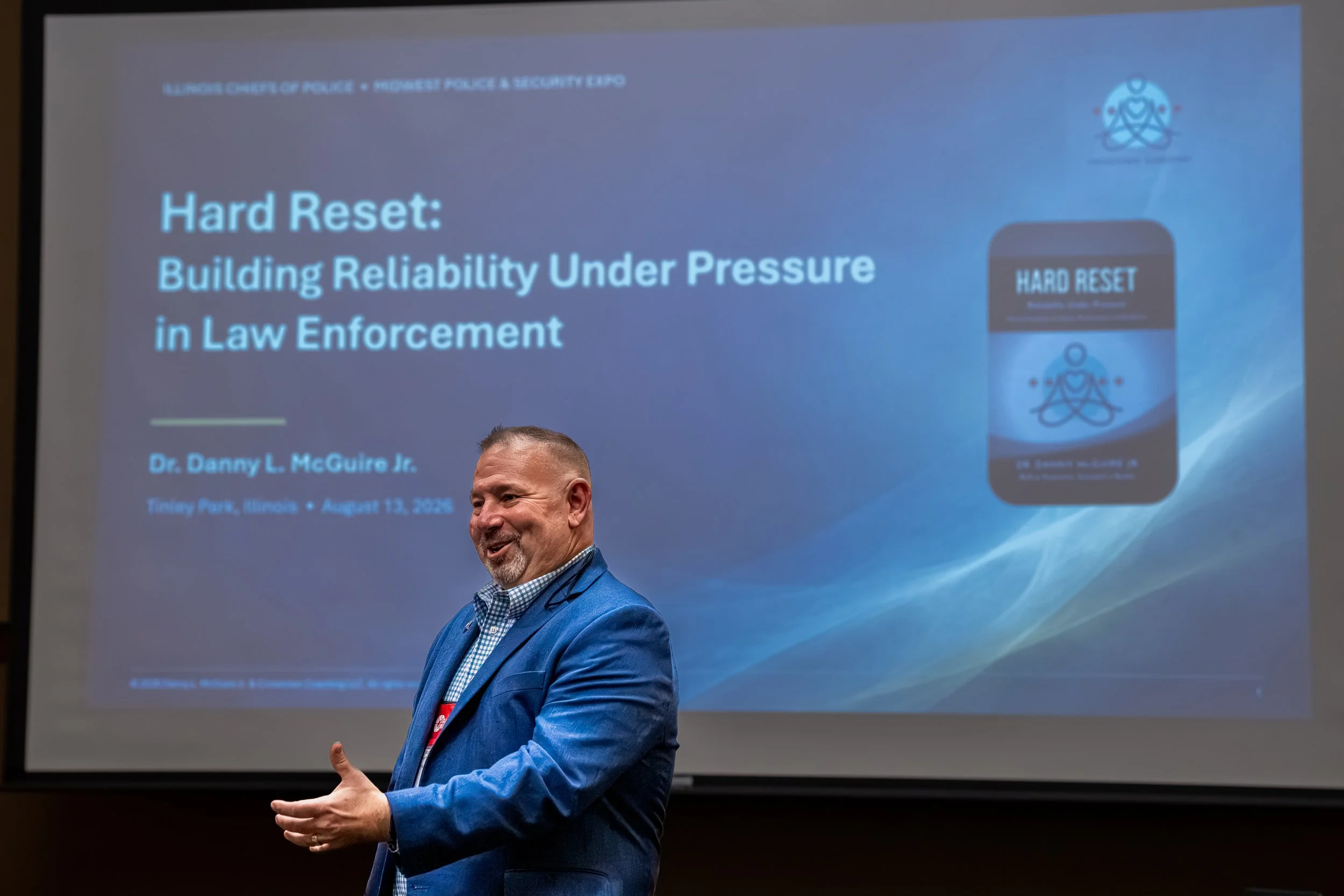 A man in a blue blazer and checkered shirt speaking at a conference with a presentation slide in the background titled "Hard Reset: Building Reliability Under Pressure in Law Enforcement." The slide features a graphic of a smartphone displaying the s