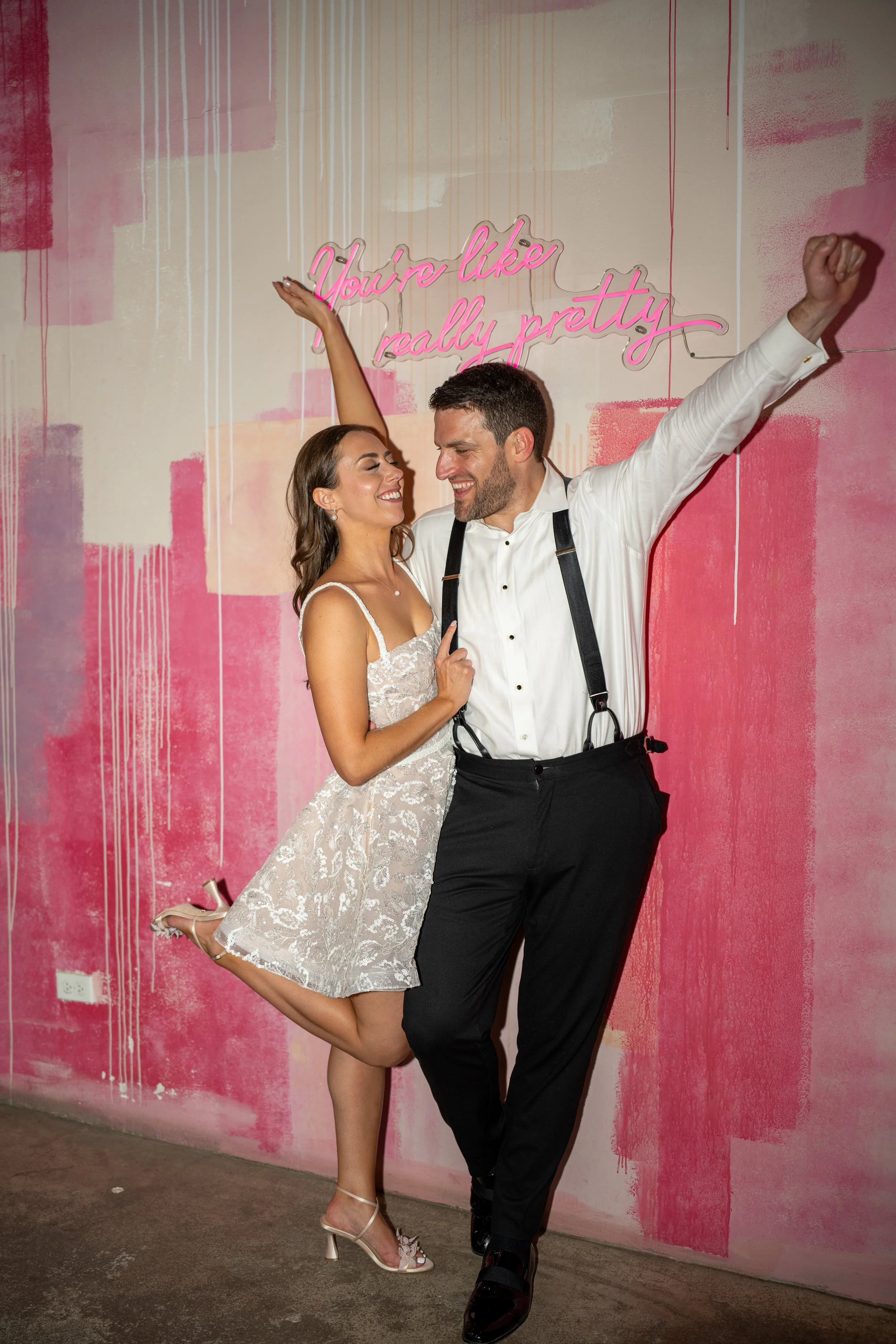 A woman in a lace dress and high heels jumping with one leg bent backward, smiling, and a man in a white shirt and black suspenders dancing with a big smile, standing against a painted pink wall with a neon sign that reads 'You're like really pretty.