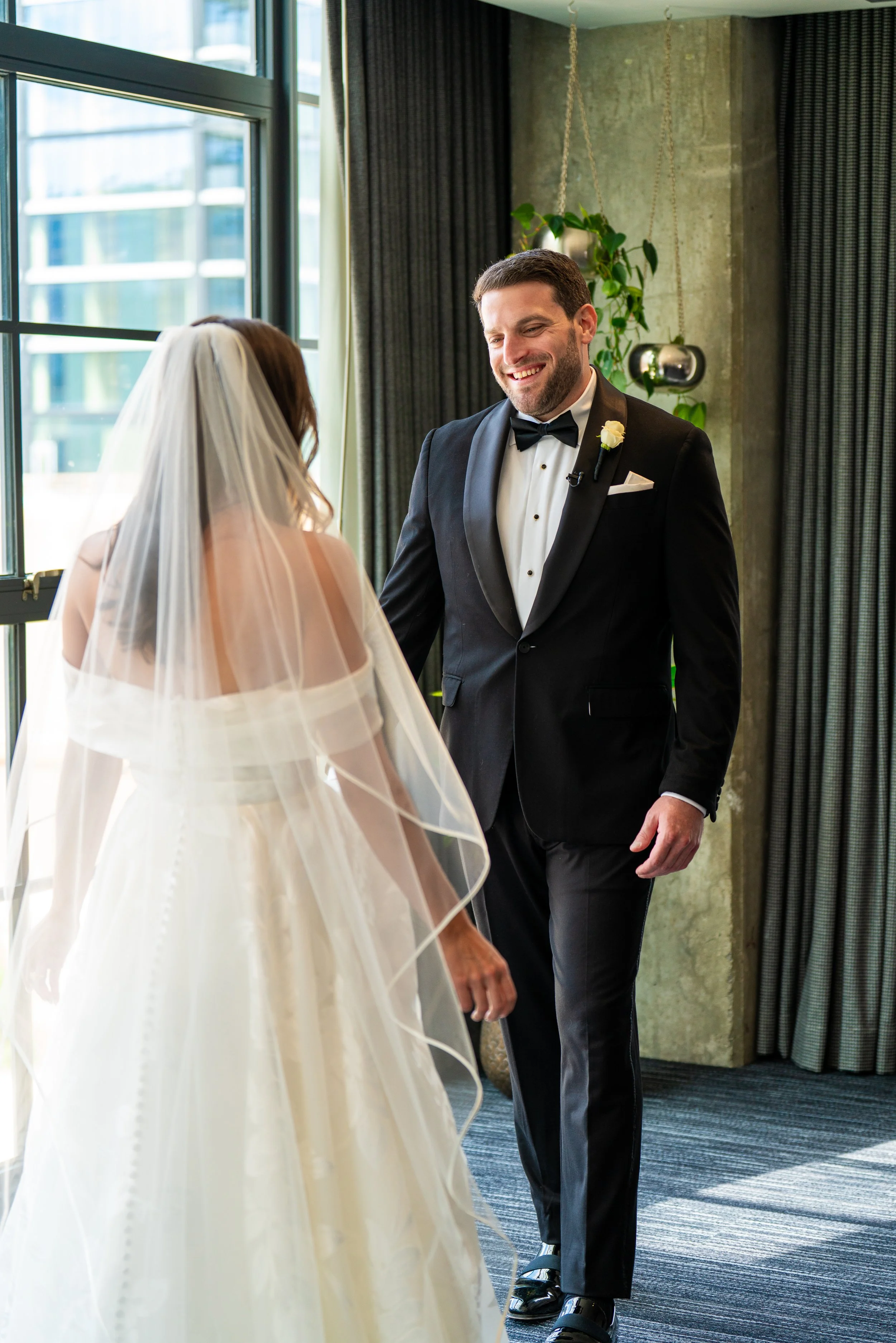 A man in a tuxedo with a bow tie, smiling, standing opposite a woman in a wedding gown with a veil, inside a modern room with large windows, curtains, and a potted plant.