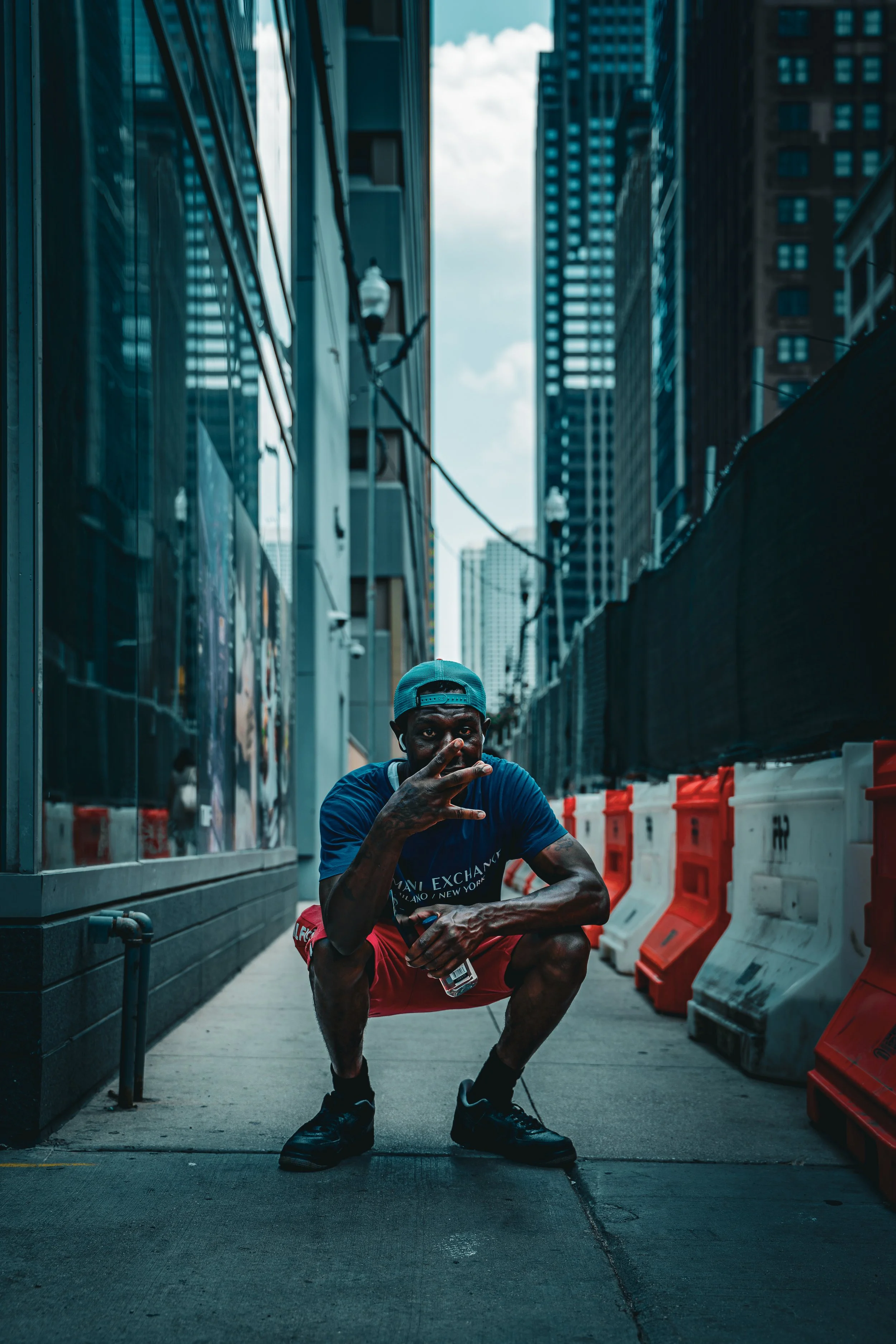A man squatting on a city sidewalk, wearing a blue cap backwards, a navy t-shirt, red shorts, and black sneakers. He is holding a water bottle and making a peace sign with his right hand. The scene is set between tall buildings with colorful barriers
