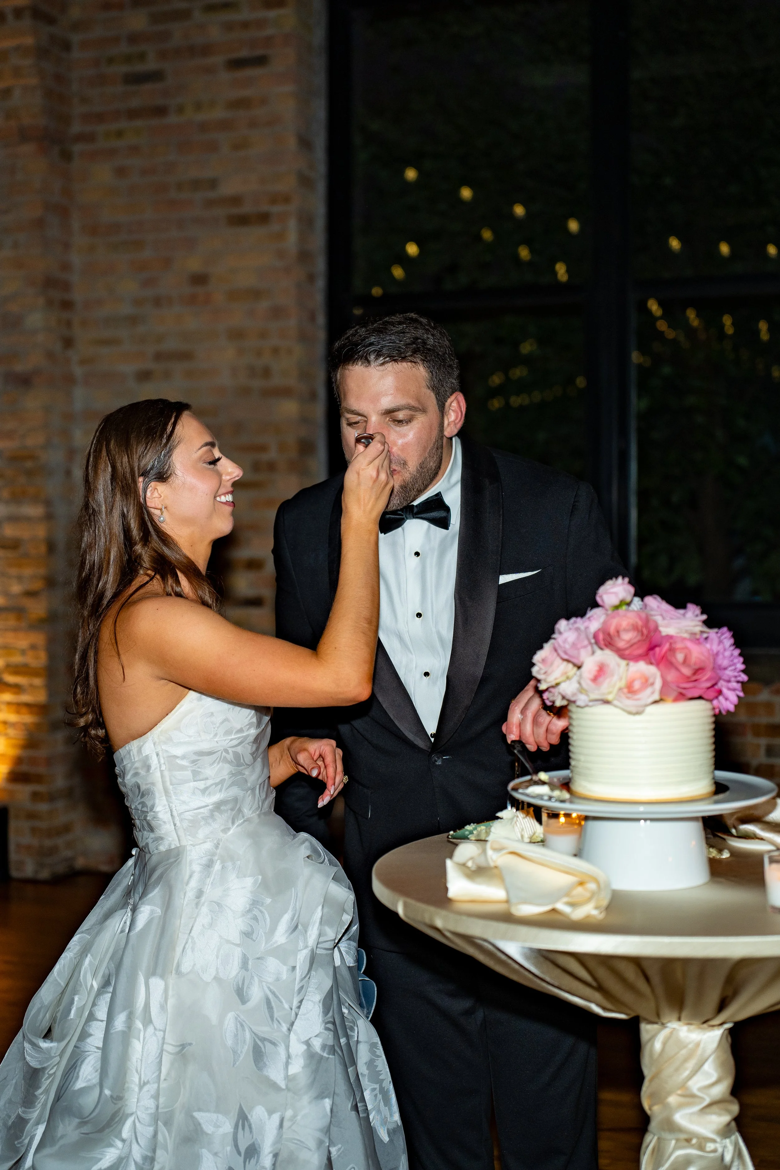 A bride and groom at their wedding cake, with the bride feeding the groom a piece of cake. The bride is smiling and wearing a white wedding gown, and the groom in a black tuxedo with a bow tie. The wedding cake is decorated with pink and white flower