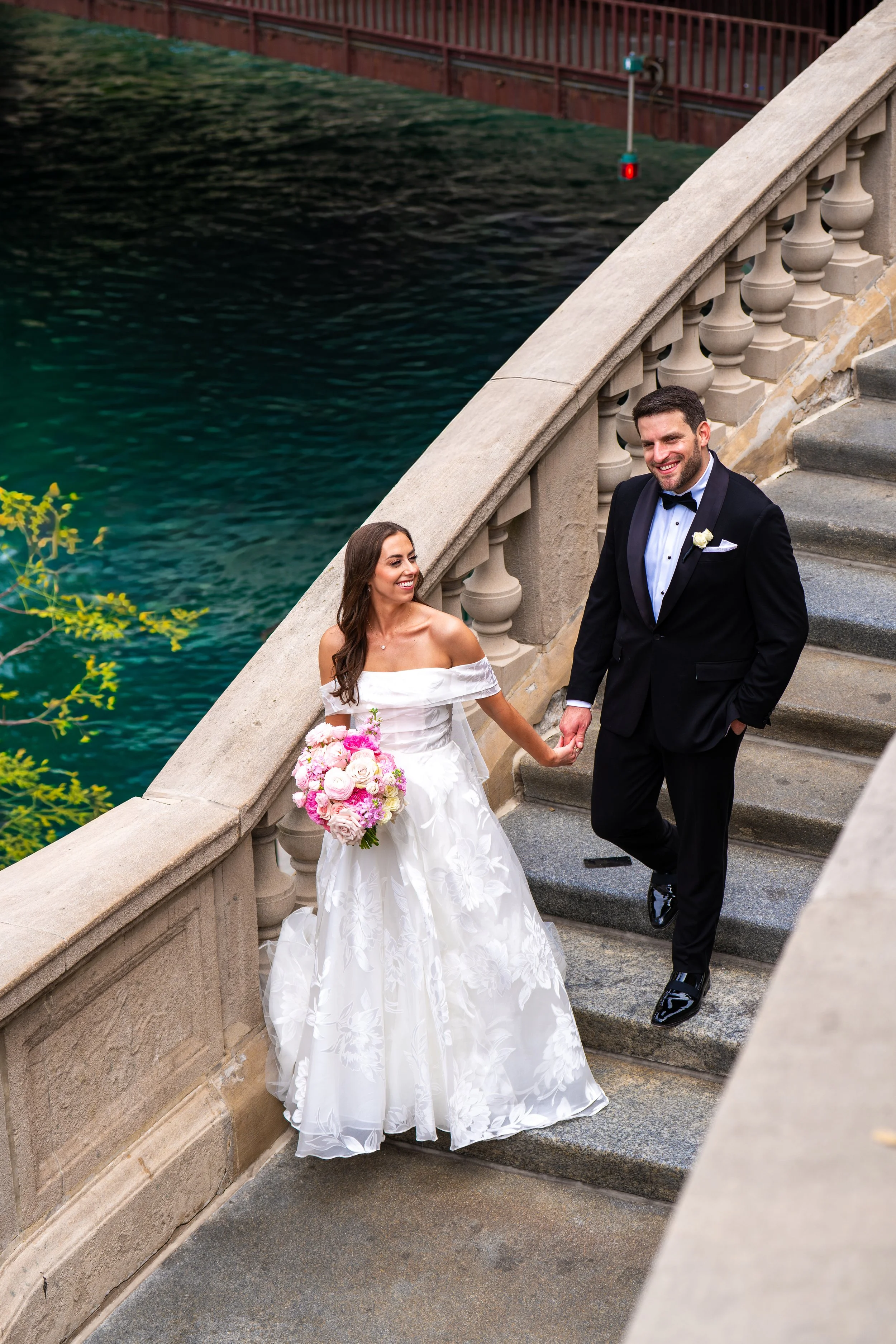 A newlywed couple holding hands and smiling on a stone staircase beside a body of water. The bride is wearing a white strapless wedding dress and holding a bouquet of pink and white flowers. The groom is dressed in a black tuxedo with a black bow tie