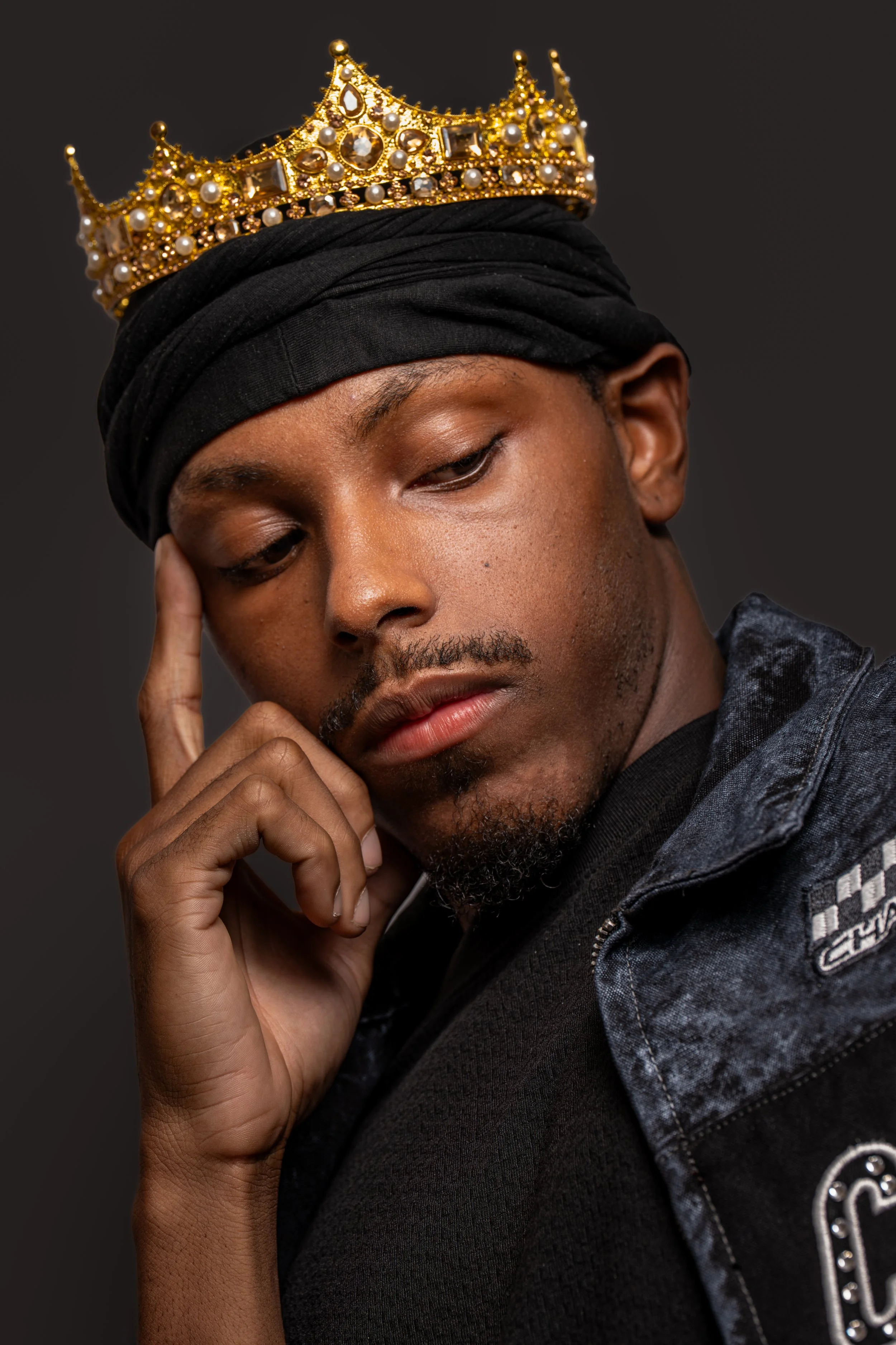 A young man wearing a black head wrap and a black jacket with a logo, with a golden crown adorned with pearls and gemstones on his head, touching his face with his right hand, against a dark background.
