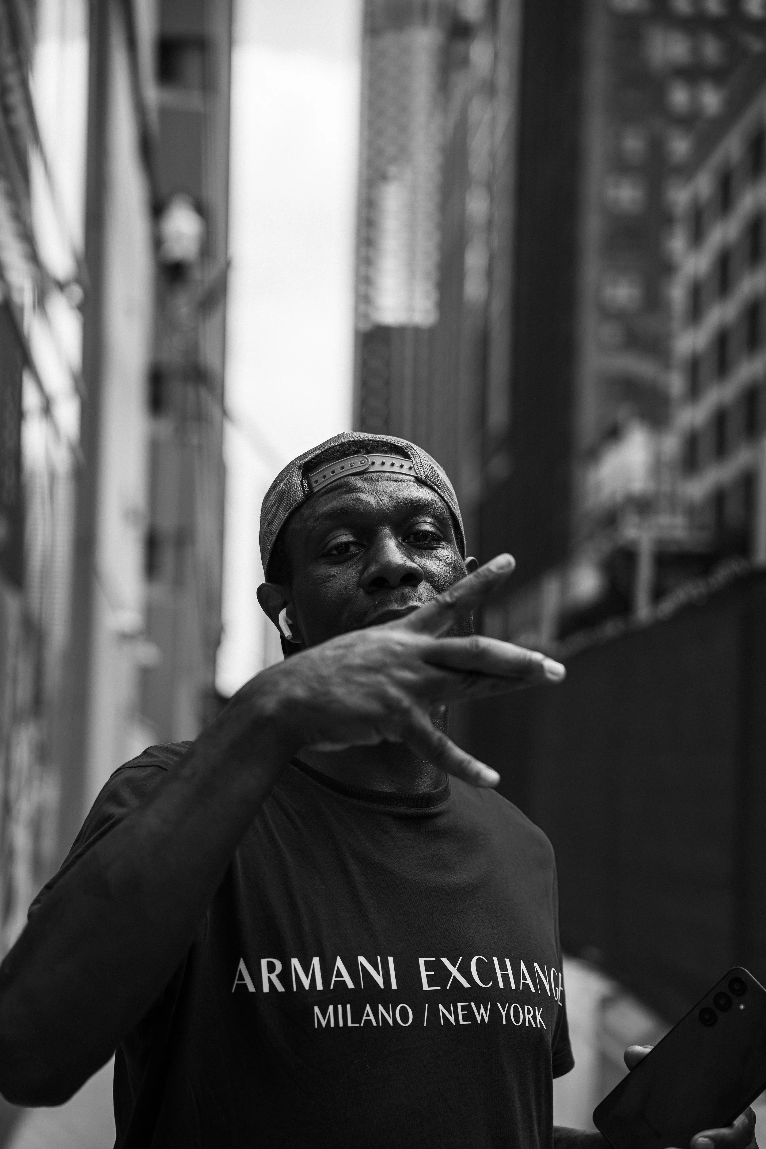A man wearing a baseball cap and a black Armani Exchange t-shirt, holding a tablet in his left hand, making a gesture with his right hand, standing outdoors in an urban setting in front of tall buildings.