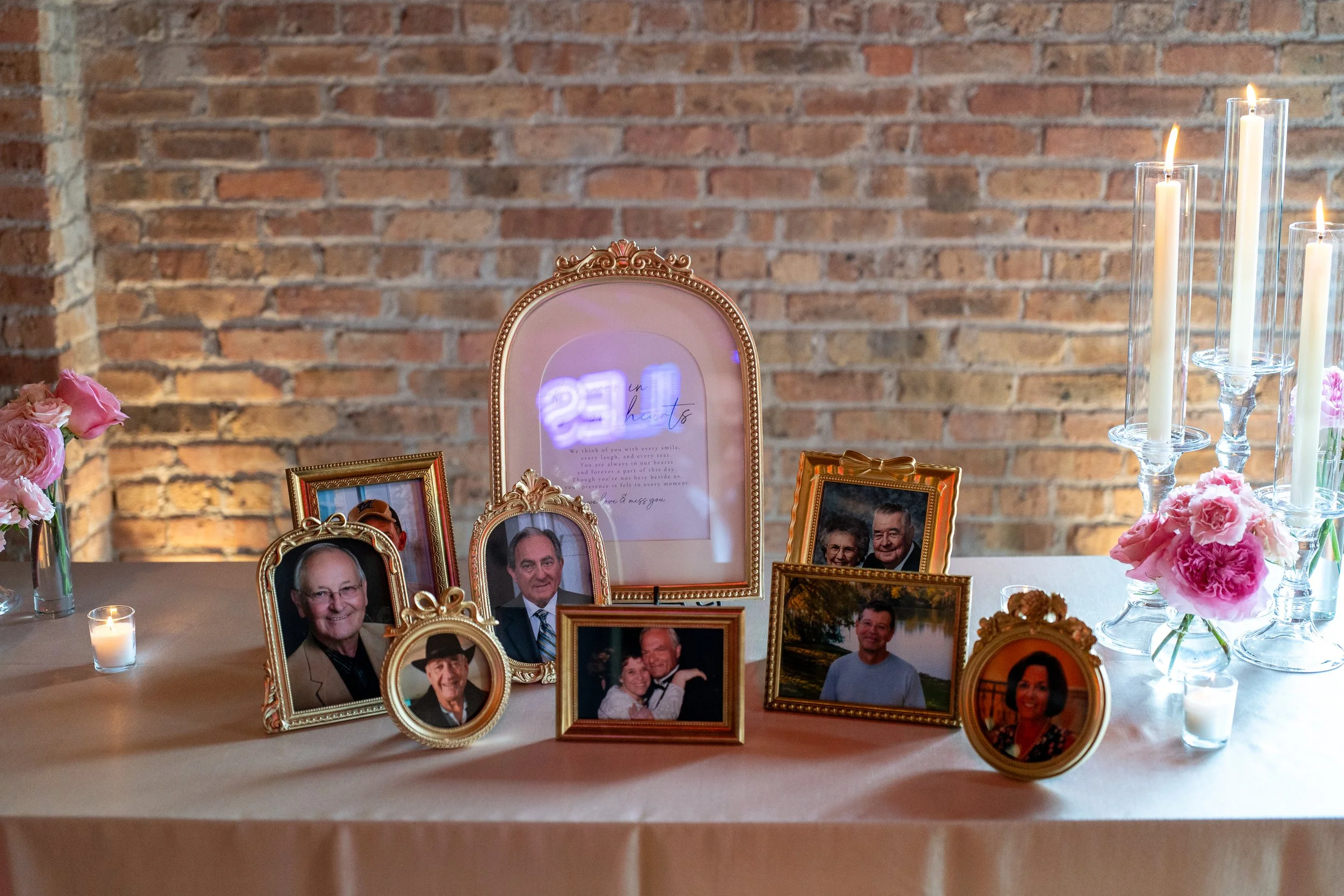 Memorial display of framed photographs of various people, including elderly individuals and couples, arranged on a table with pink flowers and candles against a brick wall backdrop.