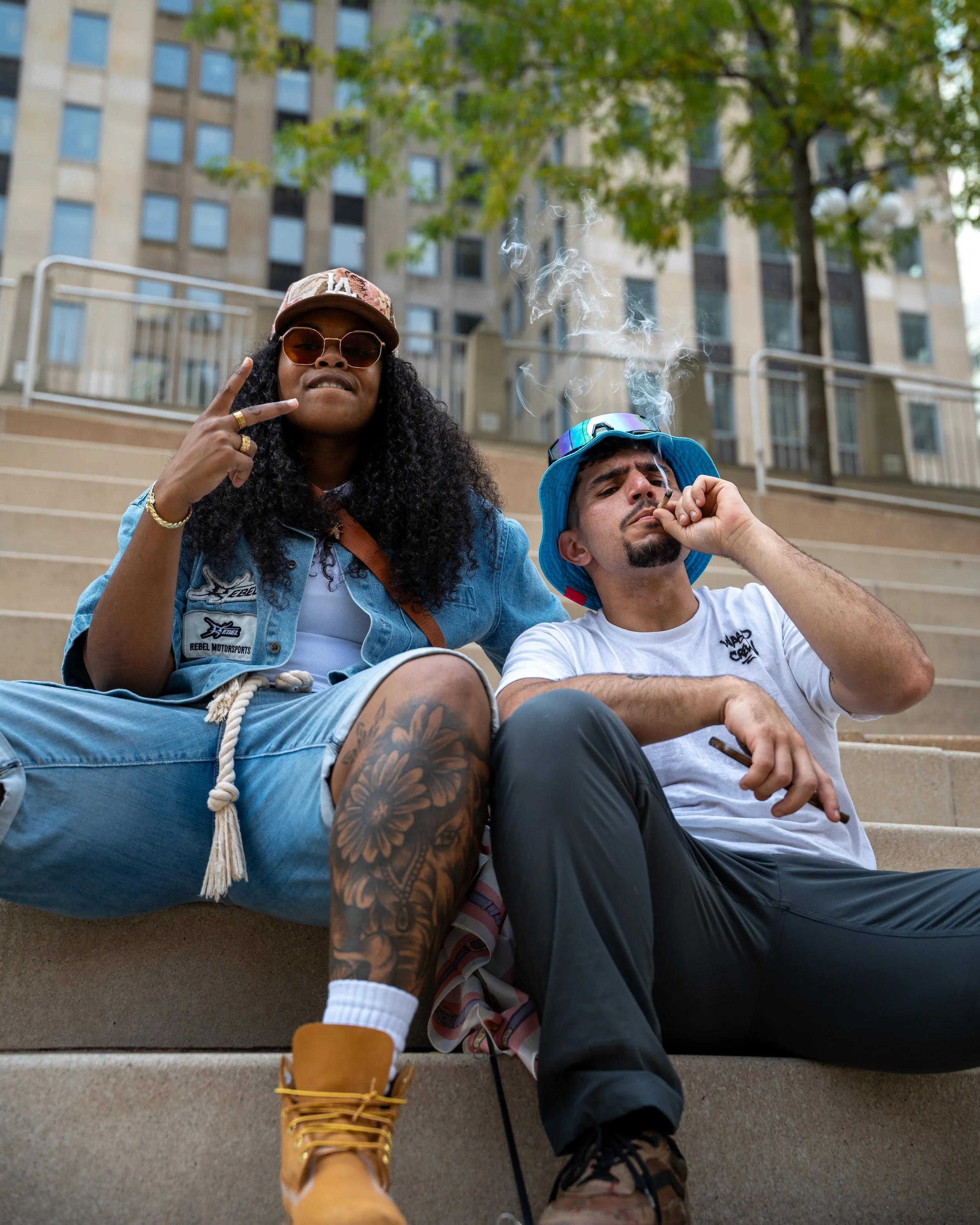 Two people sitting on outdoor stairs in an urban setting, one woman and one man, both smoking, with a background of buildings and trees.
