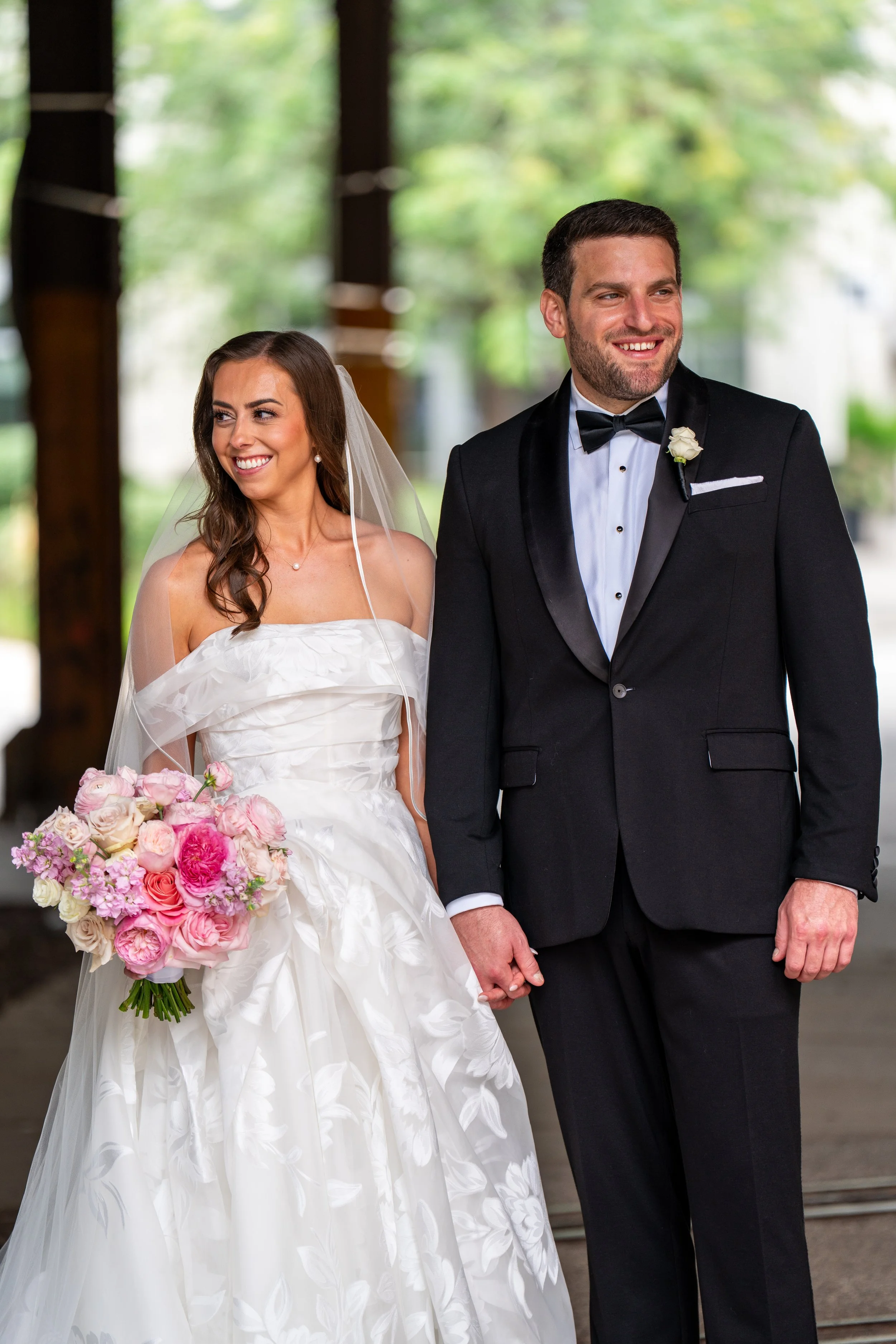 A bride and groom dressed in wedding attire holding hands and smiling, standing outdoors with a blurred greenery background.