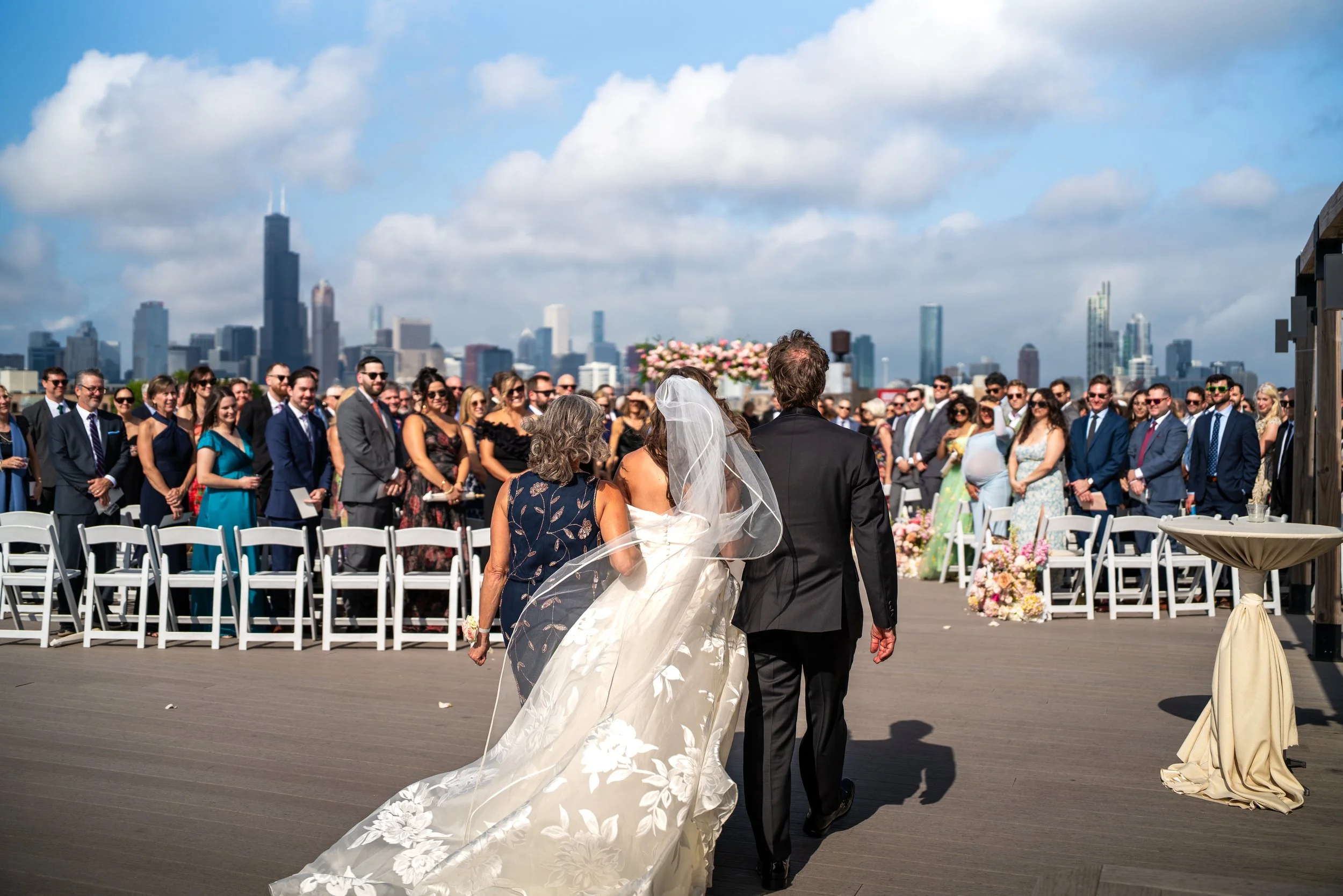 A bride in a white wedding gown and veil walking arm-in-arm with a groom in a black suit, at a wedding ceremony outdoors on a rooftop with city skyline in the background.